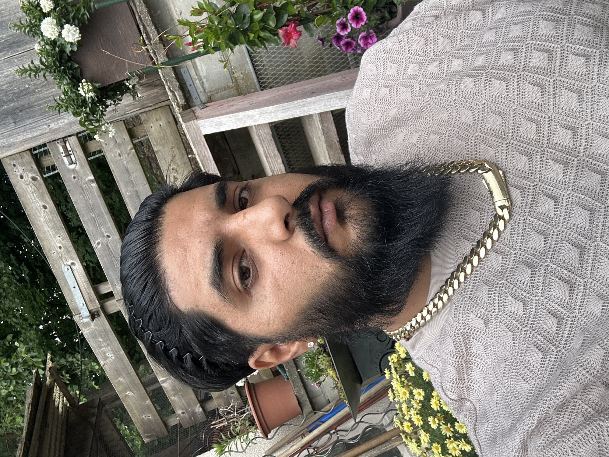 An adult male individual is depicted in a close-up, head-and-shoulders portrait, looking directly at the camera in an outdoor setting in Mainburg, Germany. He has dark, slicked-back hair, secured on his left side by a black wavy metal hairband. He features a full, dark beard and mustache, along with a subtle nose ring in his left nostril and a small, clear stud earring in his left earlobe. He is wearing a light tan or beige long-sleeved knitted sweater with a textured, geometric pattern resembling interlocking diamonds or hexagons. Around his neck is a thick, gold-colored Cuban link chain, with its rectangular clasp prominently visible at the front.
The background is a garden or backyard environment, dominated by weathered wooden fences and various plants. On the left, behind the subject, a terracotta pot with a plant is situated on a ledge, with yellow daisy-like flowers visible in the lower foreground. On the right, a gray-brown wooden plank fence with a visible metal hinge extends across the frame. Hanging on this fence is a brown pot containing white flowering plants, and below it, pots hold a green plant with red flowers and a cluster of purple petunia-like flowers. Dark green foliage from trees or bushes forms the uppermost background. The scene is illuminated by natural, diffused daylight.