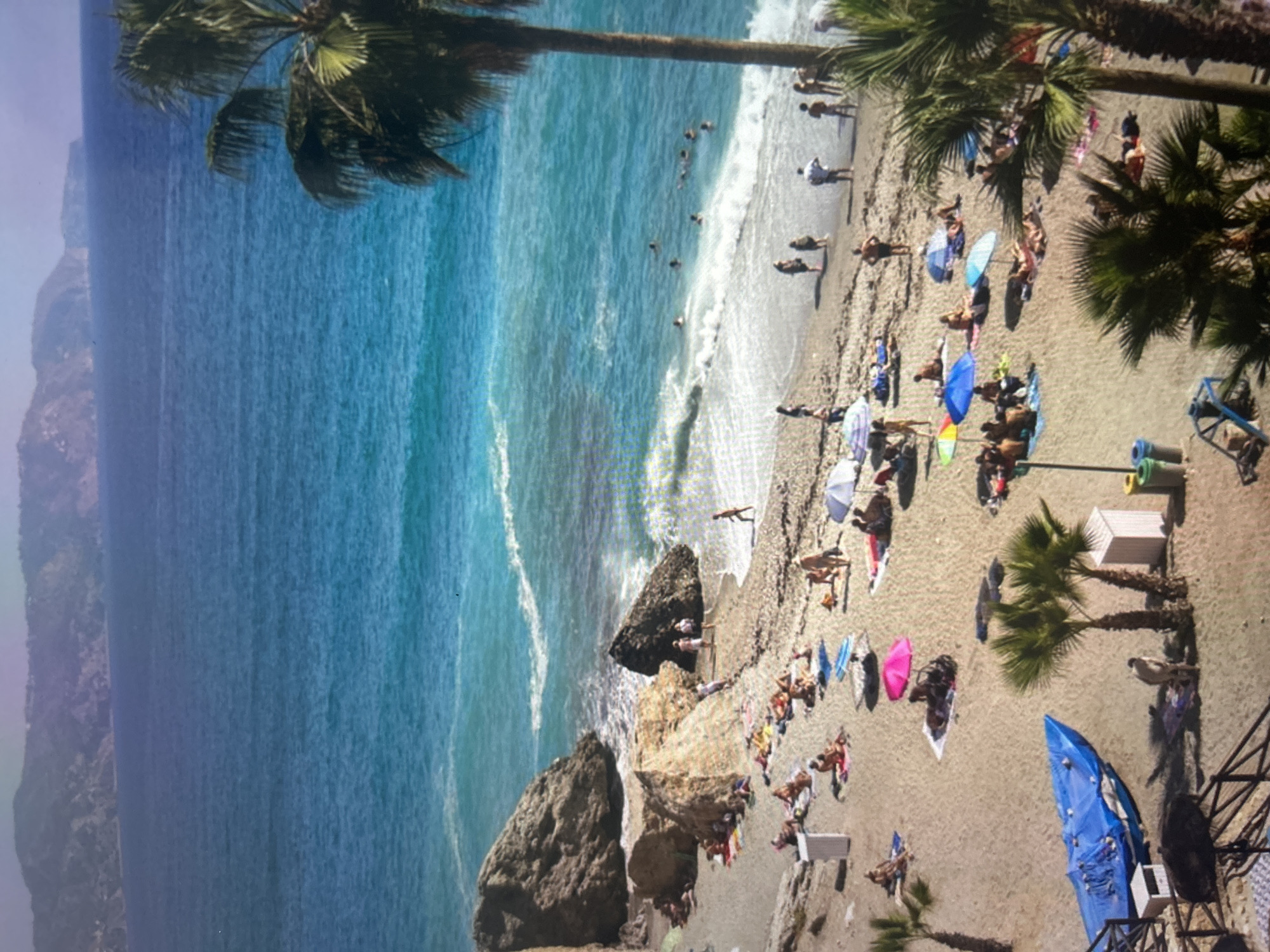 This is a high-angle view of a bustling beach on a bright, sunny day, featuring clear blue water and a crowded shoreline.
The scene is dominated by the expansive ocean on the left, displaying various shades of turquoise and deep blue, indicating clear water and depth changes. Gentle white waves lap at the shore, and several people are visible swimming, wading, or standing in the refreshing water. Closer to the beach, large, dark rock formations jut out from the water and rest on the sand, adding natural elements to the coastal landscape. In the far background, hazy, light-colored mountains or cliffs rise along the coastline.
On the right side of the image, the sandy and pebbled beach is teeming with activity. Numerous people are enjoying the day, with many sunbathing on towels, sitting on loungers, or strolling along the shore. Colorful beach umbrellas in blue, pink, white, and striped designs provide shade for sunbathers. Several lush palm trees are scattered along the beachfront, casting shadows and adding a tropical feel. One large palm tree extends diagonally across the top of the frame. Toward the bottom right, a prominent blue boat or kayak rests on the sand, near a small, white structure and what appears to be a railing or part of a beach facility. Other beach accessories like trash cans are also visible.
The clear lighting and distinct shadows indicate it is daytime, likely during the peak hours of sunlight. The atmosphere suggests a popular vacation destination, with people enjoying leisurely activities in warm, pleasant weather. No discernible text is visible within the image.