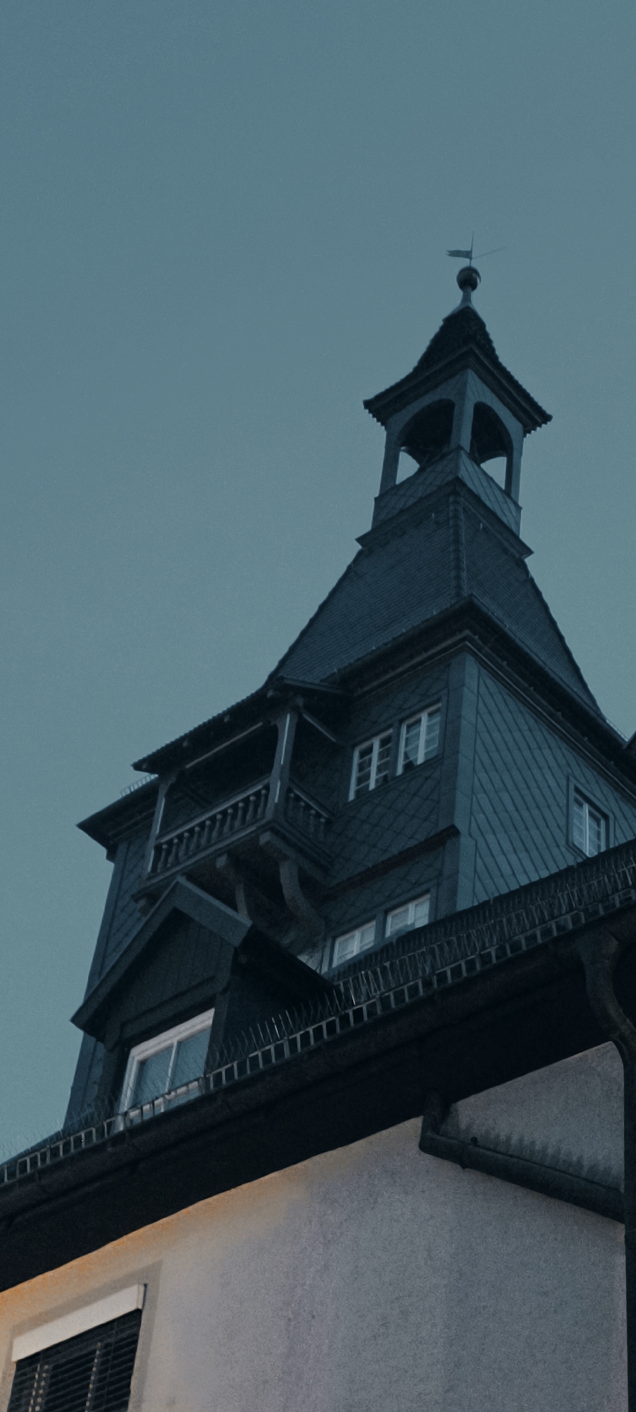 The image shows a close-up, low-angle view of the spire and upper facade of a dark-colored building, likely a historic structure, against a muted teal sky. The building's architecture features a prominent, multi-tiered dark roof, possibly slate or tile, culminating in a tower with arched openings and a weather vane. A balcony with a wooden railing extends from the main structure. Several windows with white frames are visible, some with multiple panes. The lower portion of the image reveals a lighter-colored, textured wall of another building, with a window and dark blinds or shutters. The overall impression is of an old building, possibly in a town or city, captured during twilight or a heavily overcast day.
This image is of the Althöfer building in Winnenden, Germany, located at Marktstraße 18. The building is known for its distinctive tower and historical significance within the city.