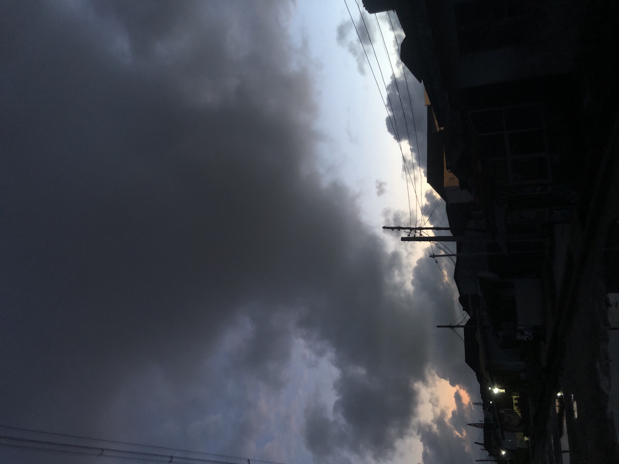 The image depicts a dimly lit street scene at dusk in Ado, Nigeria. The sky is filled with dramatic, dark, and heavy clouds, suggesting an impending storm or the aftermath of rain. The overall lighting is low, with the sky showing a gradient from dark grey overhead to a lighter, pale blue with hints of orange near the horizon on the left.
In the foreground, the ground appears wet, with puddles reflecting the faint light. Silhouettes of several low-rise buildings line the street. These buildings are dark and indistinct, with some appearing to be houses or small shops. There are visible utility poles and electrical wires crisscrossing the scene, typical of urban environments. Some of the buildings have lights on, casting a faint glow, but the majority of the structures remain in shadow. There are no clearly discernible individuals or specific actions taking place, presenting a static scene of an urban street at twilight under an overcast sky.