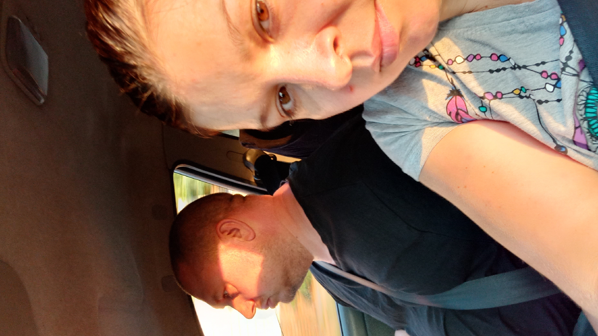 This is a portrait-oriented, close-up selfie taken from inside a vehicle, likely a car, with two individuals visible. The person taking the selfie is in the foreground, facing the camera, with their head and upper torso prominently displayed. This individual has light skin, brown eyes, and light brown hair, which is pulled back. They are wearing a light gray t-shirt with a colorful beaded necklace design printed on it, featuring feathers and beads in shades of pink, blue, yellow, and white. There's a hint of makeup on their face, and a small reddish mark is visible on their left cheekbone.
To the left and slightly behind, a man is visible in profile, facing towards the left side of the frame. He has light skin, a shaved head, and is wearing a black t-shirt. The setting sun casts a warm, orange light on his face and the interior of the car, creating strong shadows. Both individuals appear to be passengers or occupants of the vehicle, with seatbelts visible. The background shows the interior of the car, including the car ceiling and a blurred view of the outside environment through the window, suggesting motion, possibly on a road within or near the city of Kropyvnytskyi, Ukraine, given the context. The lighting suggests it is either sunrise or sunset.