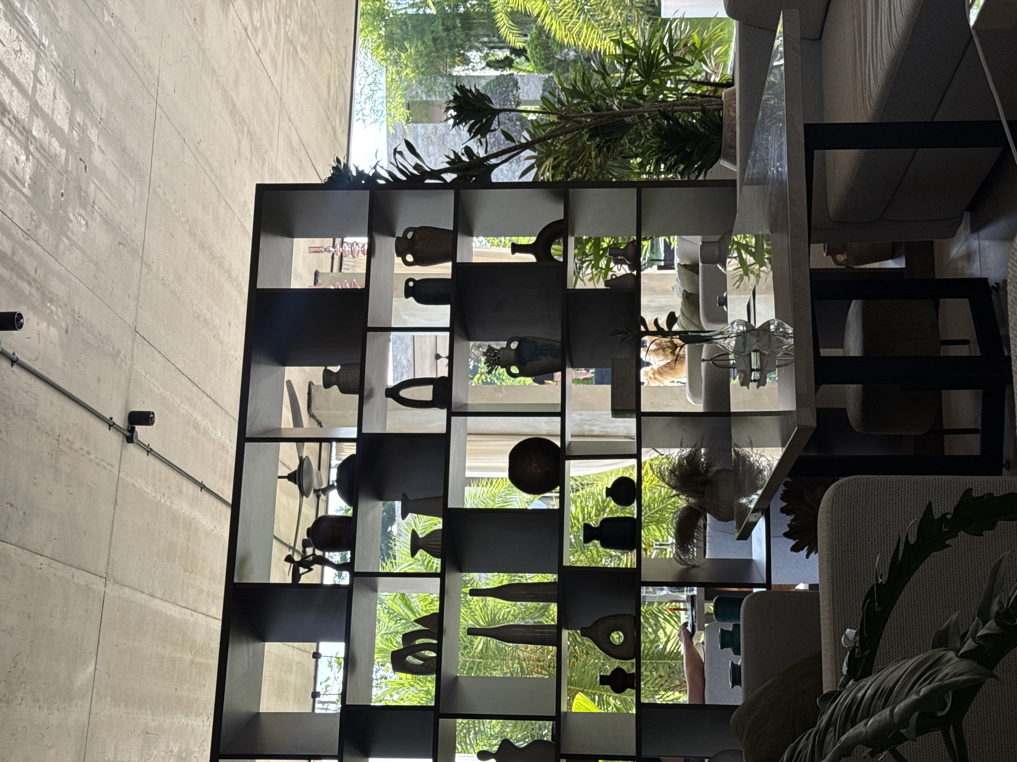 The scene depicts a shelving unit with various decorative items, likely pottery or sculptural pieces, displayed in its compartments. The shelving unit is placed in front of a large window offering a view of lush greenery, suggesting a tropical environment. Light filters through the window, casting reflections and shadows on the shelves and items.
Beyond the shelving unit, there is seating, possibly a sofa or chairs, indicating an interior space designed for relaxation. The overall aesthetic is modern and minimalist, with a focus on natural light and the incorporation of natural elements such as plants.
Given the description, the image appears to be taken in a well-designed interior space in the Ubud District of Indonesia, likely a hotel, resort, or residential property with a focus on blending indoor and outdoor elements.
