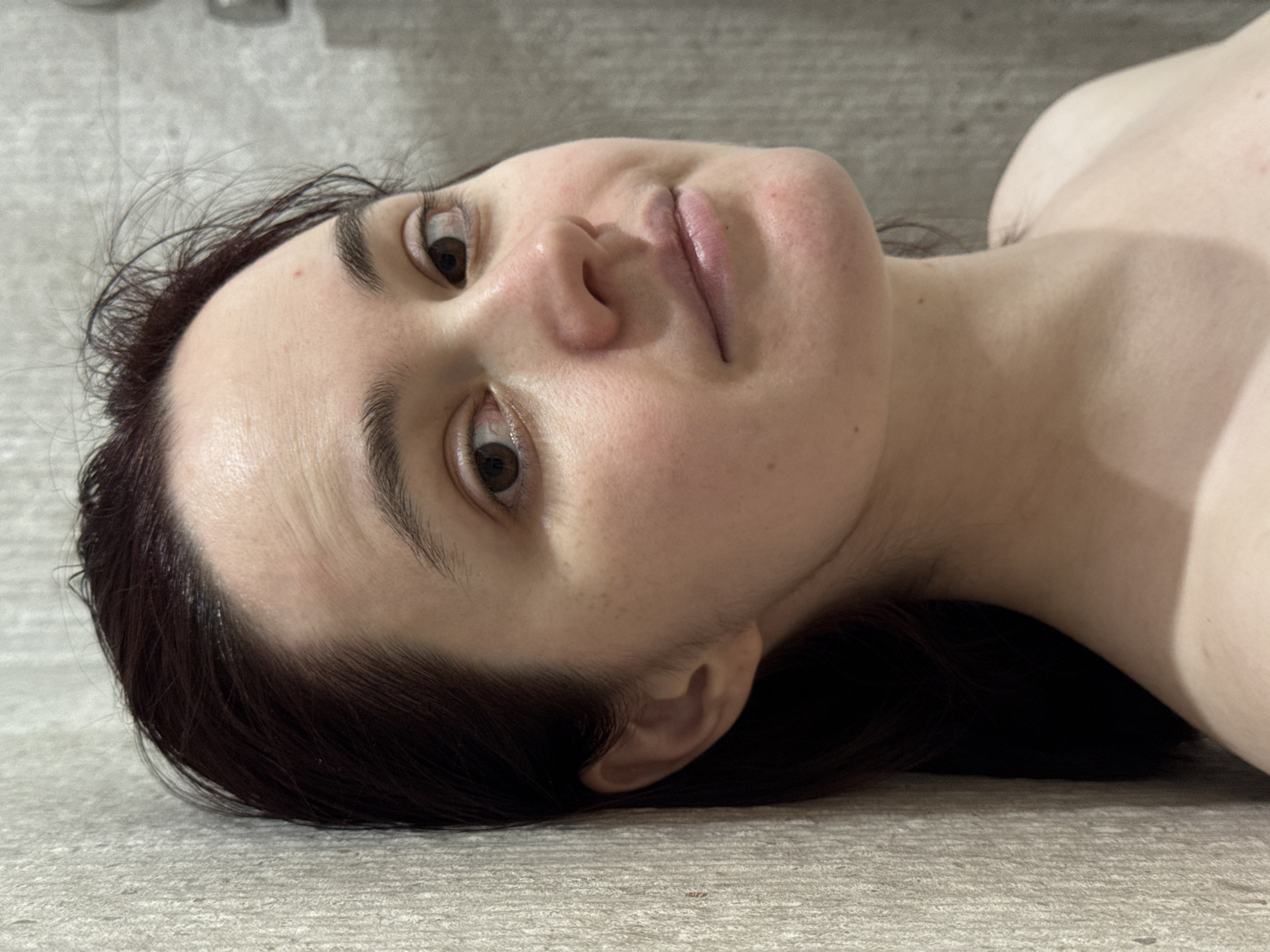 A fair-skinned person with dark brown hair is lying on a light gray tiled surface. The person's face is turned towards the camera. Their eyes are open, and they are looking at the camera. The person's left ear is visible. Part of the person's shoulder and upper chest is visible in the upper right of the frame. The location of the image is not possible to determine from the visual information.