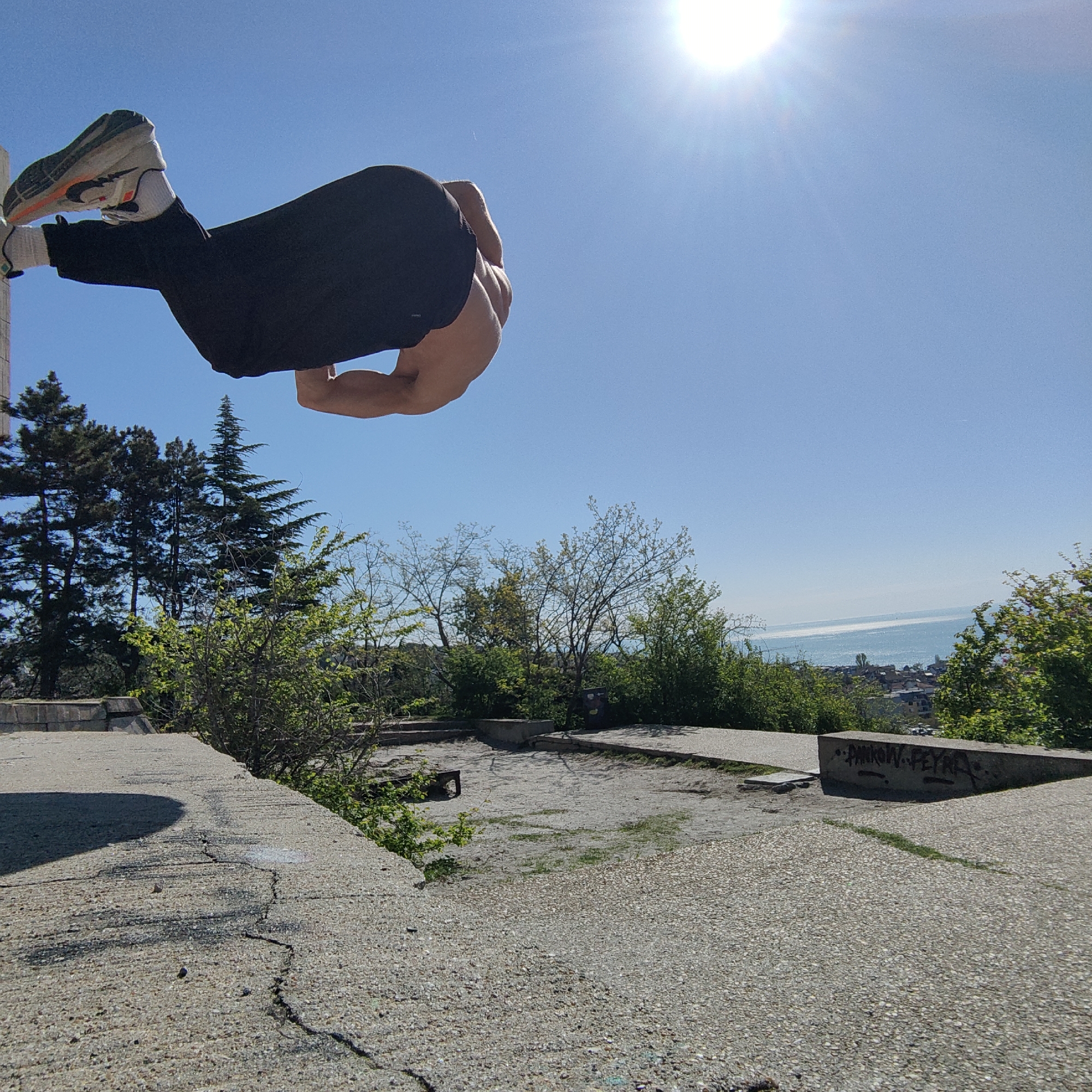 A man is in mid-air performing a backflip. He is wearing black pants, white socks, and running shoes with white, black, and orange coloring. The background includes trees, the sky, and the Black Sea. The ground is paved with concrete and shows signs of wear and cracks. Graffiti is visible on the ledge of the pavement. The location is in Varna, Bulgaria.