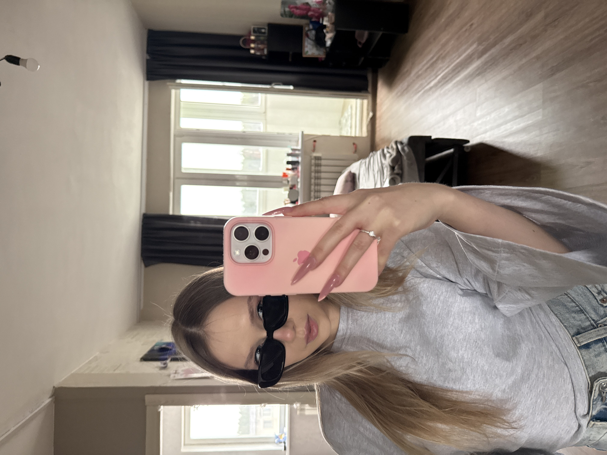 A young woman wearing sunglasses is taking a selfie indoors, using a pink smartphone. She is wearing a grey t-shirt and denim jeans. The location appears to be an apartment in the city of Chaikovsky, Russia. The apartment has dark curtains, large windows, a radiator, shelving units and light-colored wooden flooring.