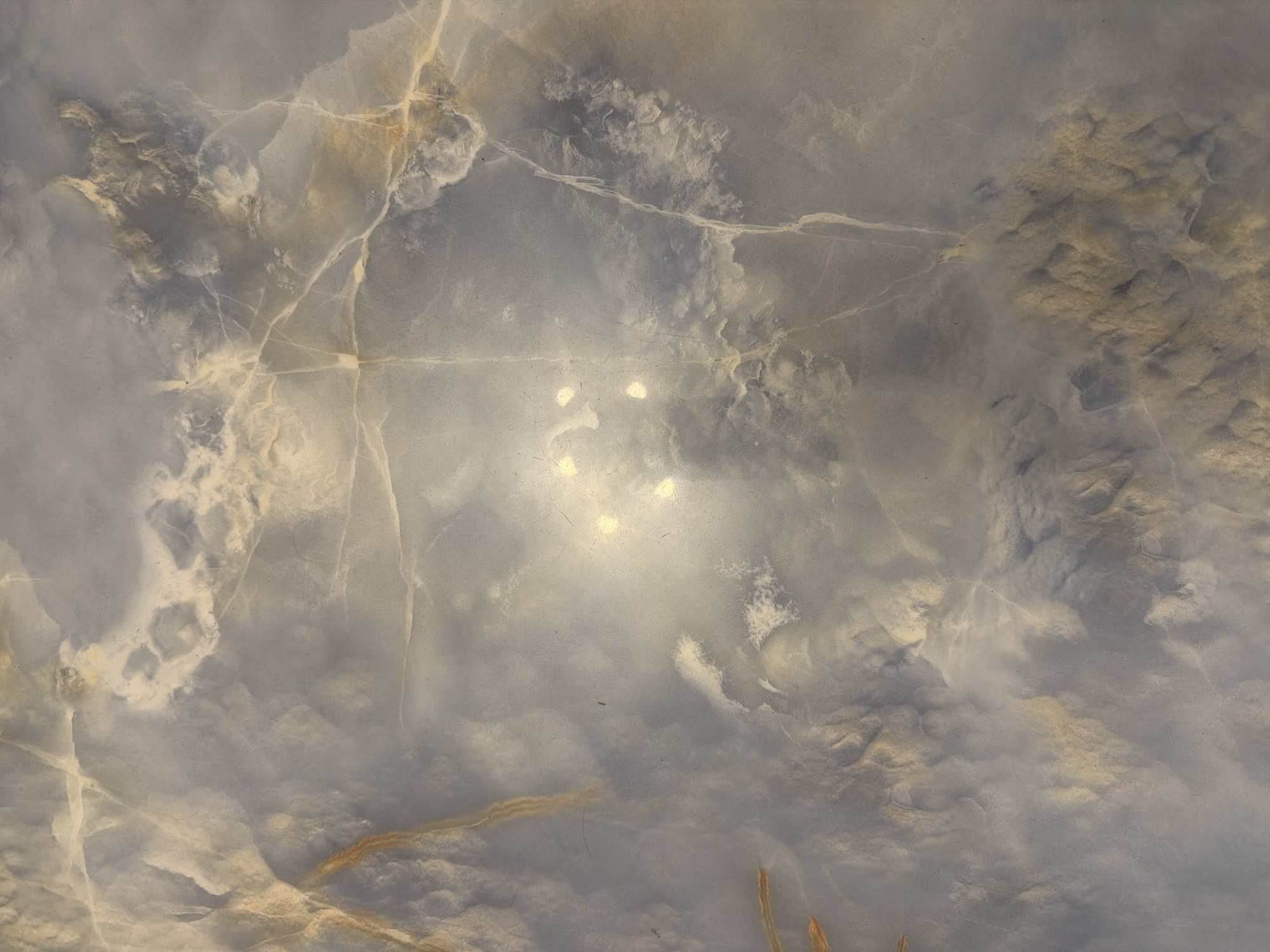 The image is a close-up, abstract shot of a polished stone surface, likely marble or onyx, with swirling patterns of grey, white, and hints of golden-brown. The texture appears smooth and reflective. There are no people, animals, or discernible objects present. The lighting creates subtle highlights and shadows, enhancing the natural veining and depth of the stone. The background is entirely composed of this textured stone surface. The location is not specified and is therefore listed as Unknown, Unknown.