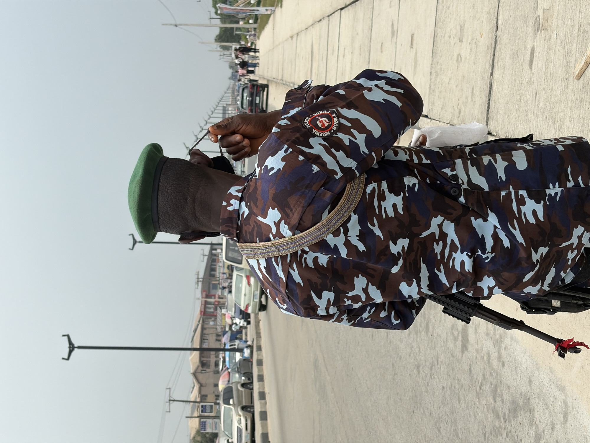 A male individual, viewed from behind, stands on a paved sidewalk in Lekki, Nigeria. He wears a dark blue, brown, and light blue camouflage uniform, topped with a green beret. A circular patch on his left sleeve identifies him with "NIGERIA POLICE FORCE MOBILE." An assault rifle, featuring a red fabric detail tied near the muzzle, is slung across his back/side. His right hand holds a pair of glasses near his ear, while a white object is tucked into his left side. To the left, a multi-lane asphalt road displays vehicular traffic, including a light-colored SUV and a silver sedan. Beyond the road, various single and multi-story buildings line the street. The right side of the image features a concrete sidewalk extending into the distance, with several pedestrians walking. Tall, black streetlights with distinctive bifurcated tops line the thoroughfare. The sky is uniformly overcast.