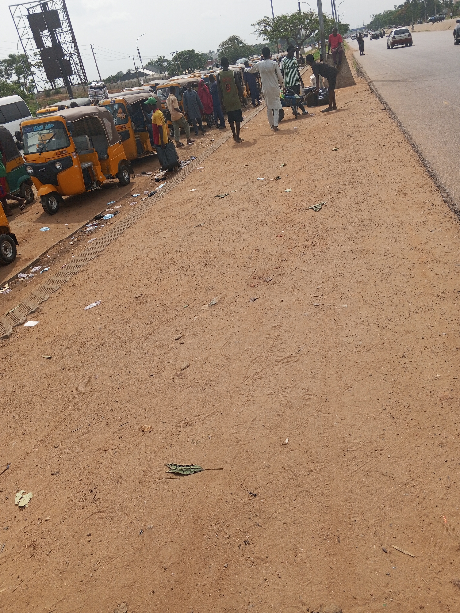 The image depicts a roadside scene in Abuja, Nigeria, characterized by the presence of yellow "keke" tricycles lined up along the shoulder of a paved road. A group of people, some carrying luggage, are standing near the tricycles, possibly waiting for transportation. Some individuals are interacting with each other, while one person is sitting on a concrete barrier along the edge of the road. The environment includes a dirt shoulder with scattered debris and tire tracks. A highway with moderate traffic is visible in the background. Overhead, there is a metal tower structure.