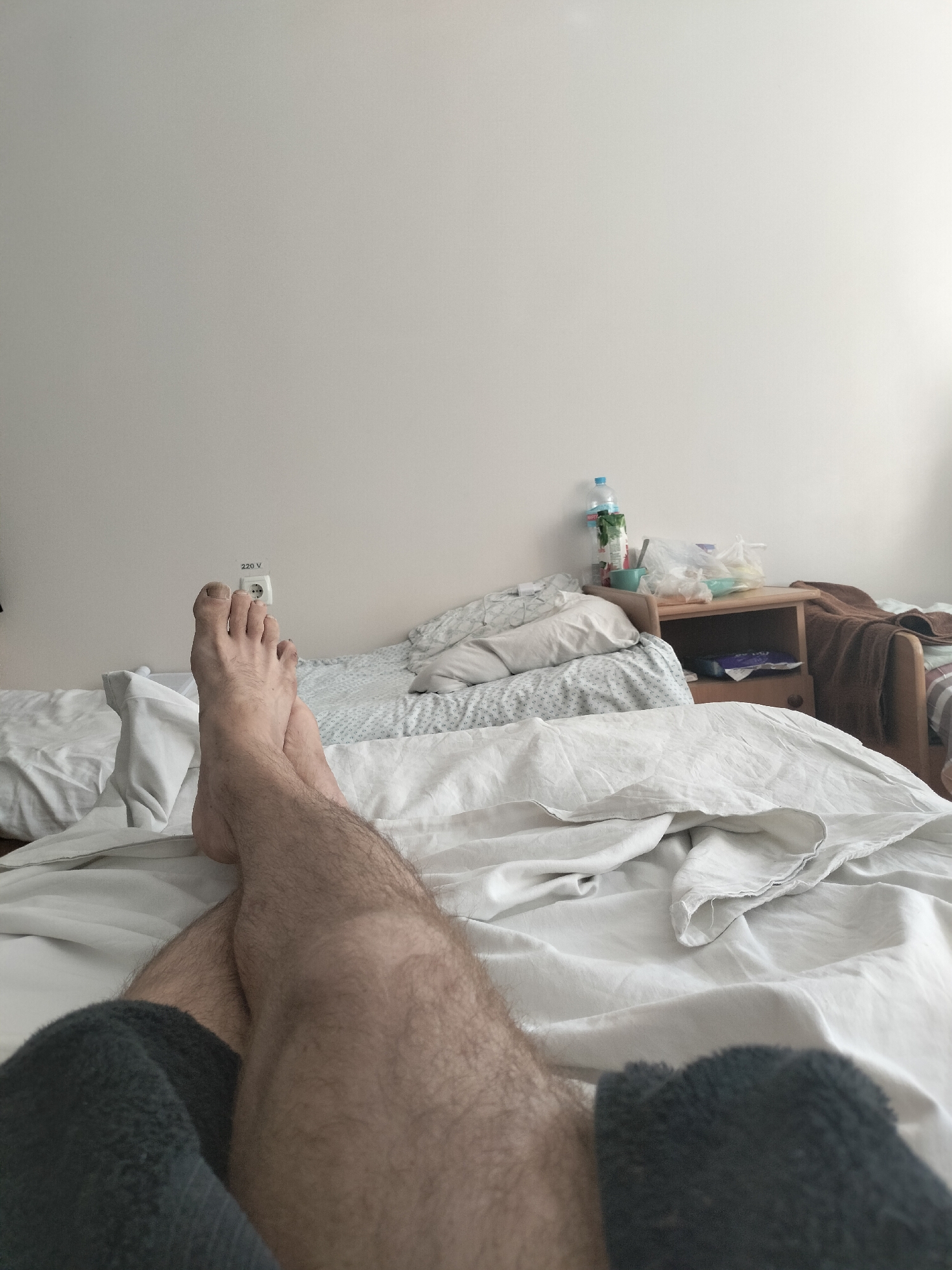 A first-person perspective shot shows a person's bare legs and feet resting on a white bedspread in a room. The person's legs are crossed at the ankles, and their feet are pointing towards the viewer. The room appears to be a bedroom, with a white wall in the background and a bedside table with some items on it. There is also a white power outlet on the wall. The lighting in the room is soft, and the overall atmosphere is relaxed.