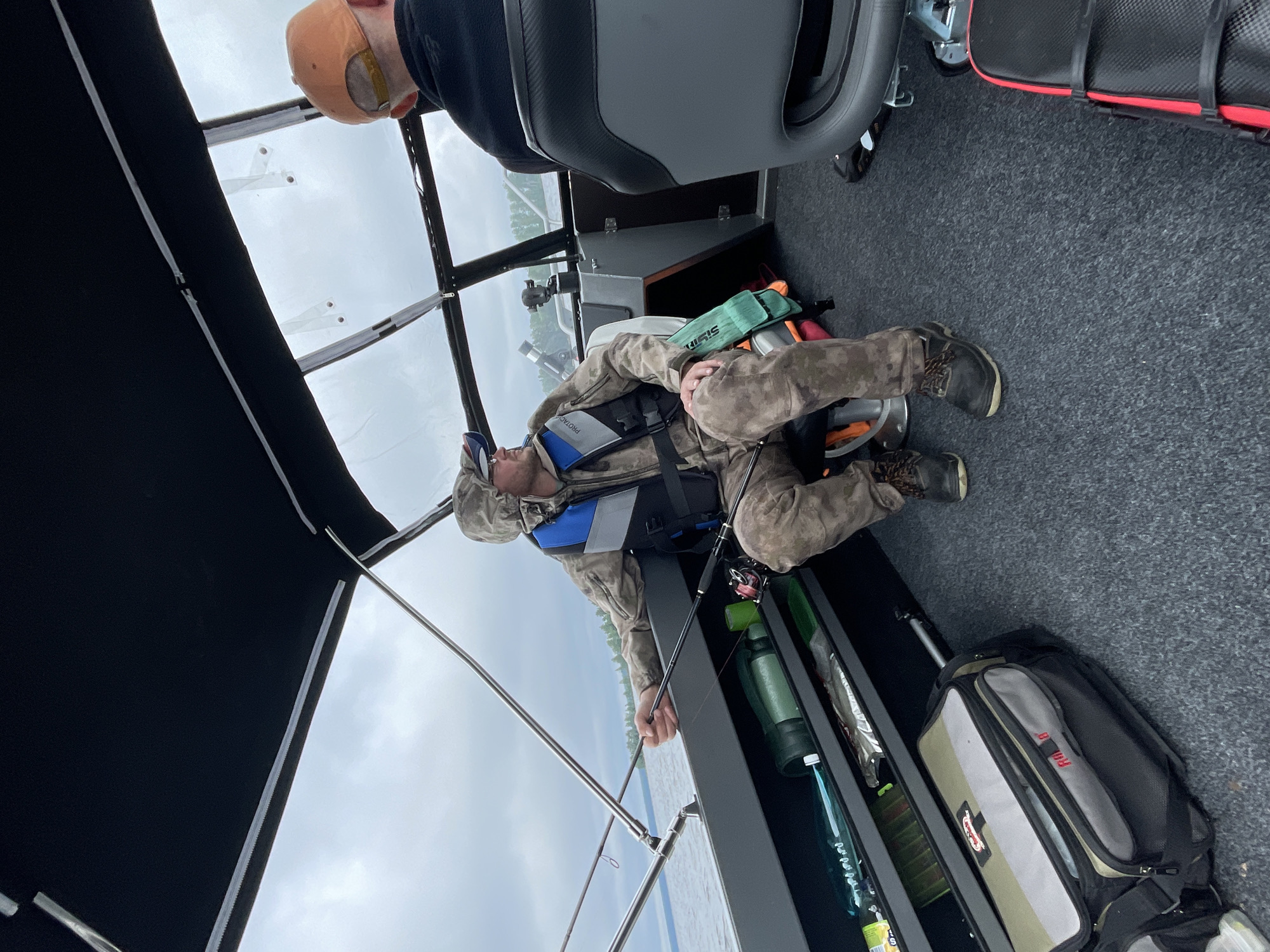 Two men are on a boat, likely in the city of Yablonovka, Russia, under a cloudy sky. The man in the foreground, wearing a camouflage jacket, camouflage pants, and a life vest, is holding a fishing rod and appears to be fishing. He is seated, facing left, and looking out at the water. Next to him are various fishing gear and a large fishing bag. The second man, wearing an orange baseball cap and a dark jacket, is seated in the background on the right side of the boat, facing away from the camera. The boat has a cabin with windows and a black canopy.
