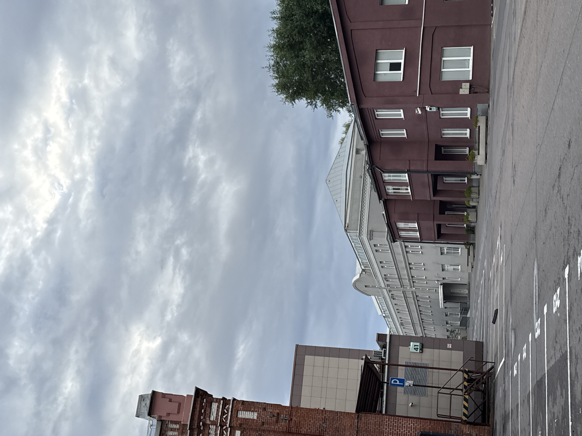 A medium shot captures an overcast sky over a commercial or industrial area in Moscow, Russia. In the foreground, a paved parking lot with white lane markings is visible. To the left, a tall brick building with decorative white detailing dominates the frame, with a section of a lighter-colored building behind it. To the right, two multi-story buildings are present: a lighter gray building with many windows and a prominent arched roof structure, and a shorter, darker red building with white-trimmed windows. A mature green tree stands behind the red building. A blue parking sign with a white "P" symbol and a sign displaying the number "41" are affixed to the lighter-colored building. Metal stairs with yellow and black hazard stripes are attached to the side of this building. A security camera is visible on the red building. The scene appears deserted.