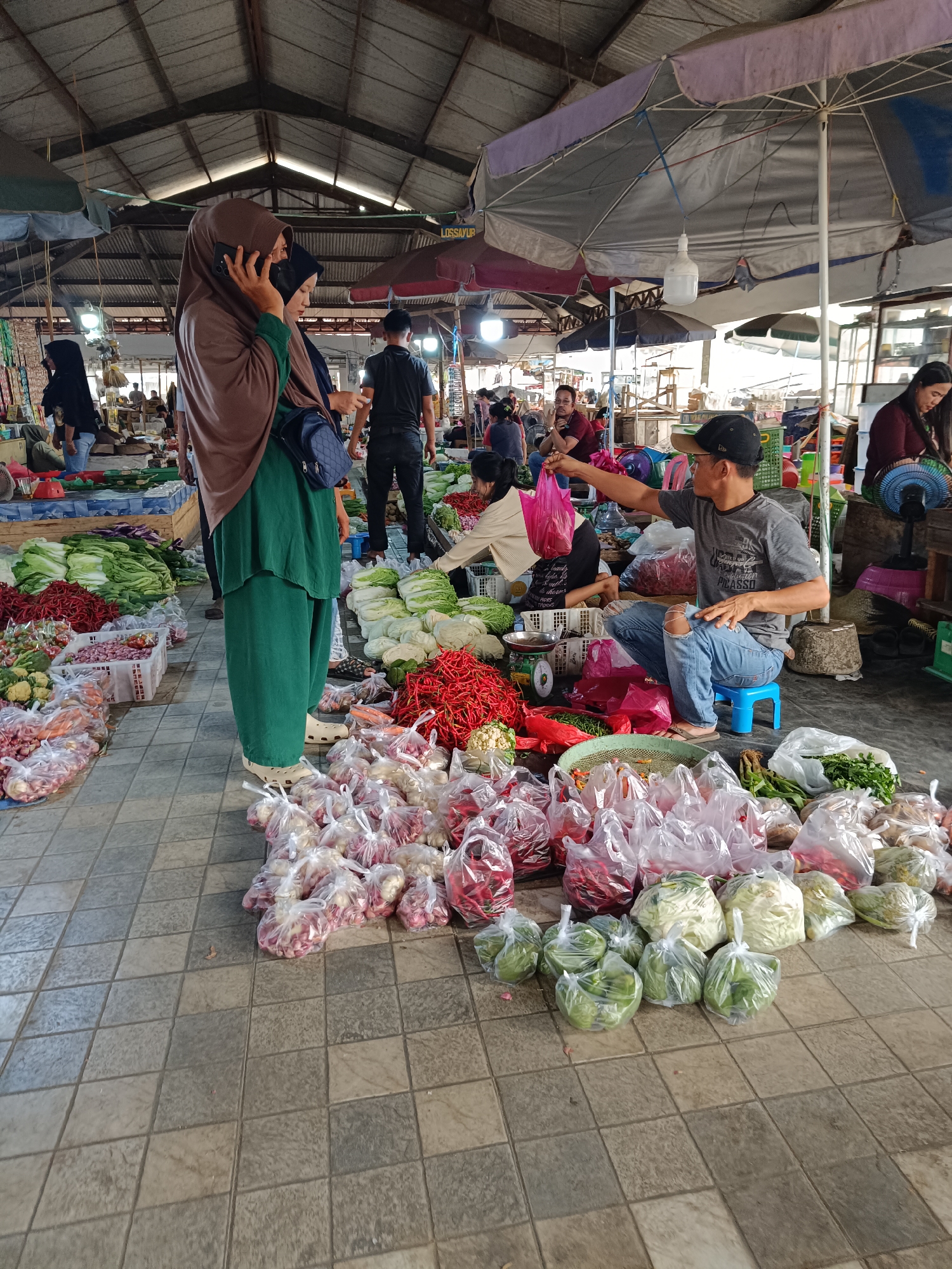 The image depicts an outdoor market scene in Kayu Agung, Indonesia, characterized by a covered stall structure with a corrugated metal roof. The ground is tiled with large, square gray tiles. A variety of fresh produce is on display, including cabbages, chili peppers, onions, and other vegetables, many of which are packaged in clear plastic bags.
In the foreground, several people are engaged in market activities. A woman wearing a brown hijab and green clothing is standing and talking on a mobile phone, with another person partially visible behind her. To the right, a man wearing a black baseball cap, glasses, and a gray t-shirt sits on a blue stool, interacting with produce and possibly a customer. He is holding up a pink plastic bag. Behind him, other individuals are visible in the background, some tending to stalls or moving through the market.
The market appears busy, with multiple stalls and a range of goods. Umbrellas provide shade over some sections of the market. The overall atmosphere suggests a typical day at a local Indonesian marketplace.