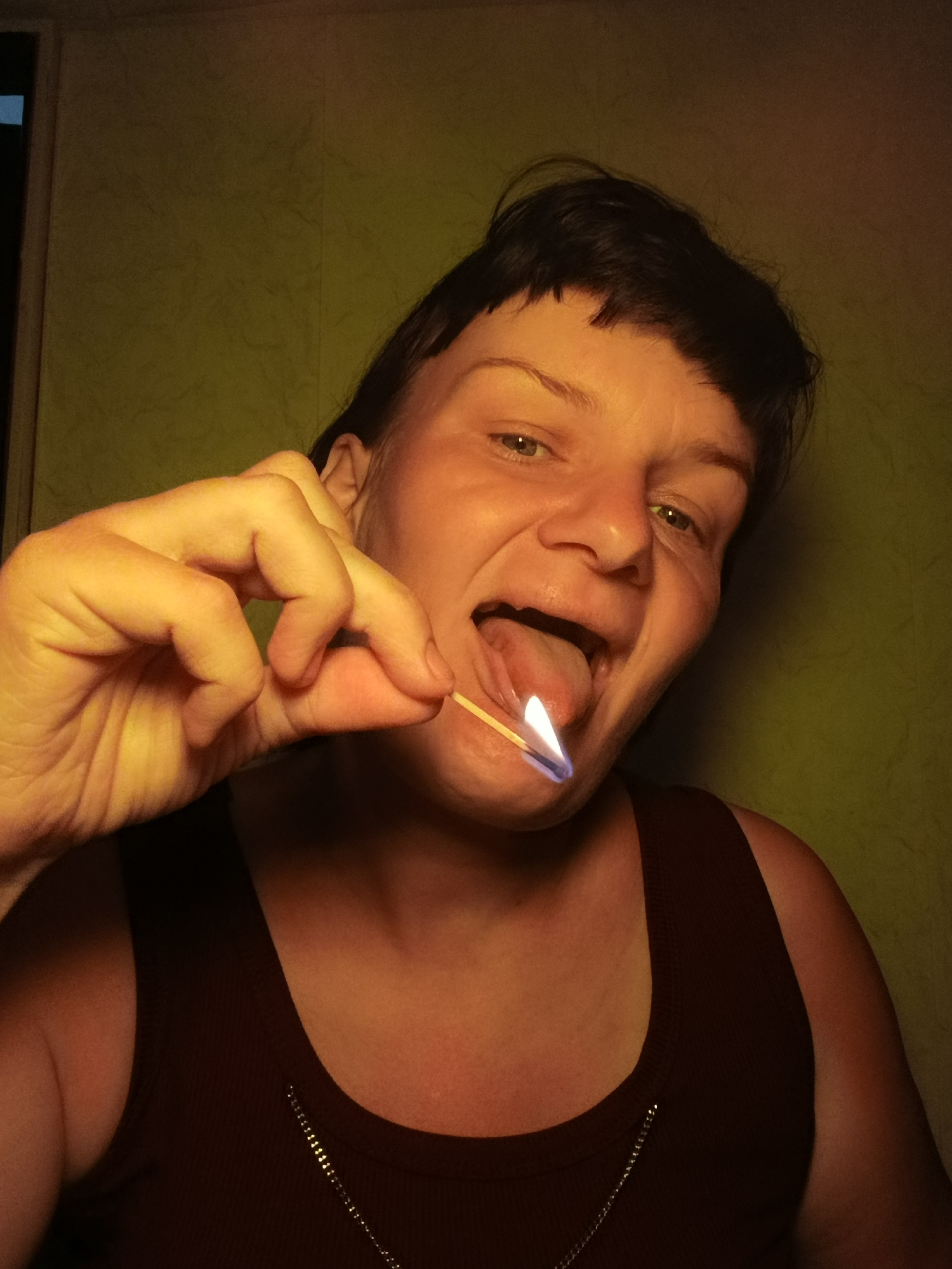 A woman is holding a lit match to her tongue, while protruding it from her mouth. She is wearing a dark tank top and has short dark hair. The background is a light green wall. There is a doorway in the left corner of the image. The location is unknown.