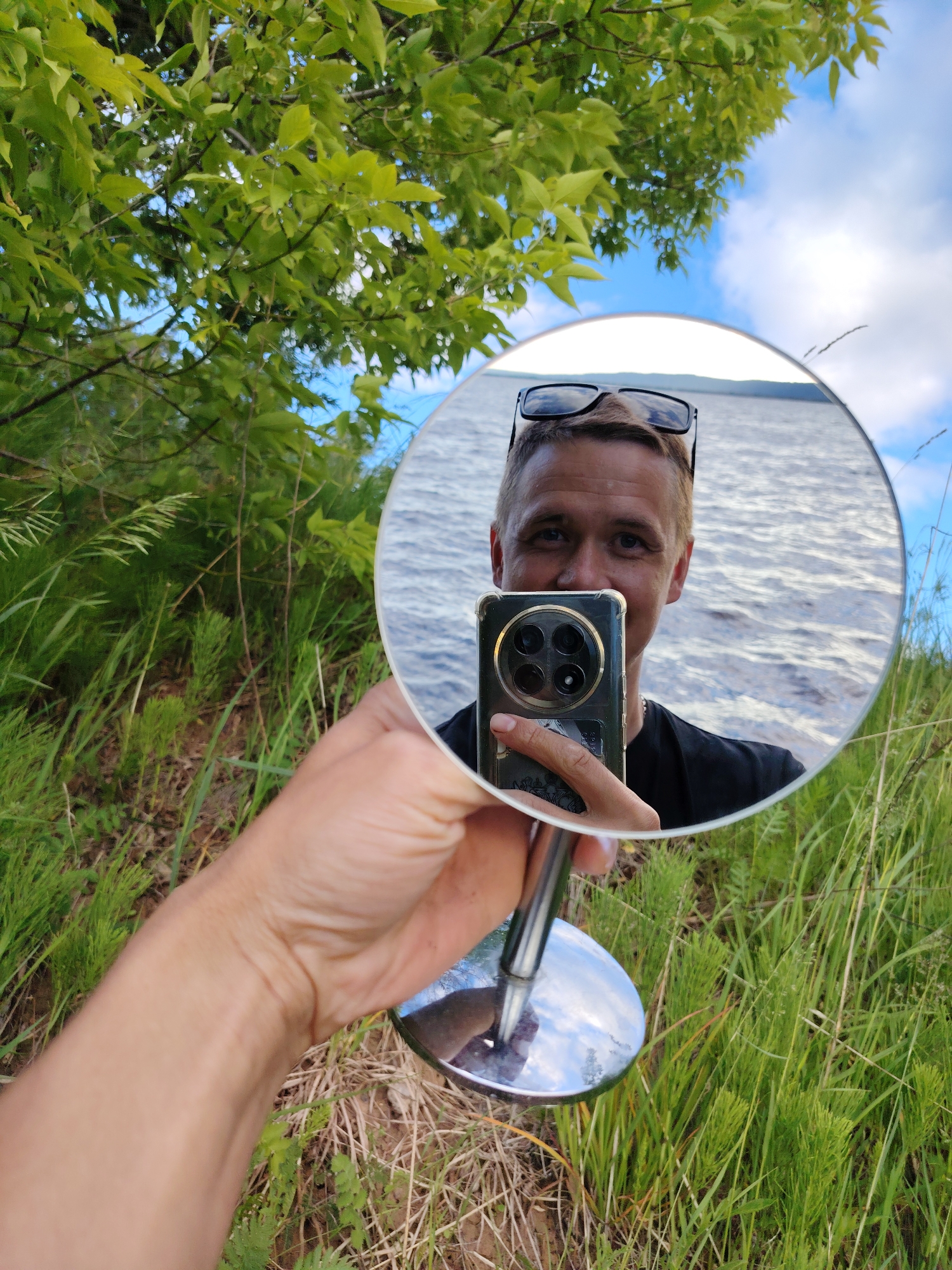 A person is taking a mirror selfie outdoors. The mirror is held by a hand in the foreground, and it reflects the person's face and upper body. They are wearing sunglasses pushed up on their head and a black t-shirt. Behind the person, a body of water is visible, with ripples on the surface, suggesting it is a lake or river. The environment is lush and green, with trees and tall grass surrounding the scene. The sky is partly cloudy, with a clear blue sky peeking through. The image appears to be taken in a natural setting, possibly a park or waterfront area in Chaikovsky, Russia, given the context. The overall atmosphere is casual and relaxed.