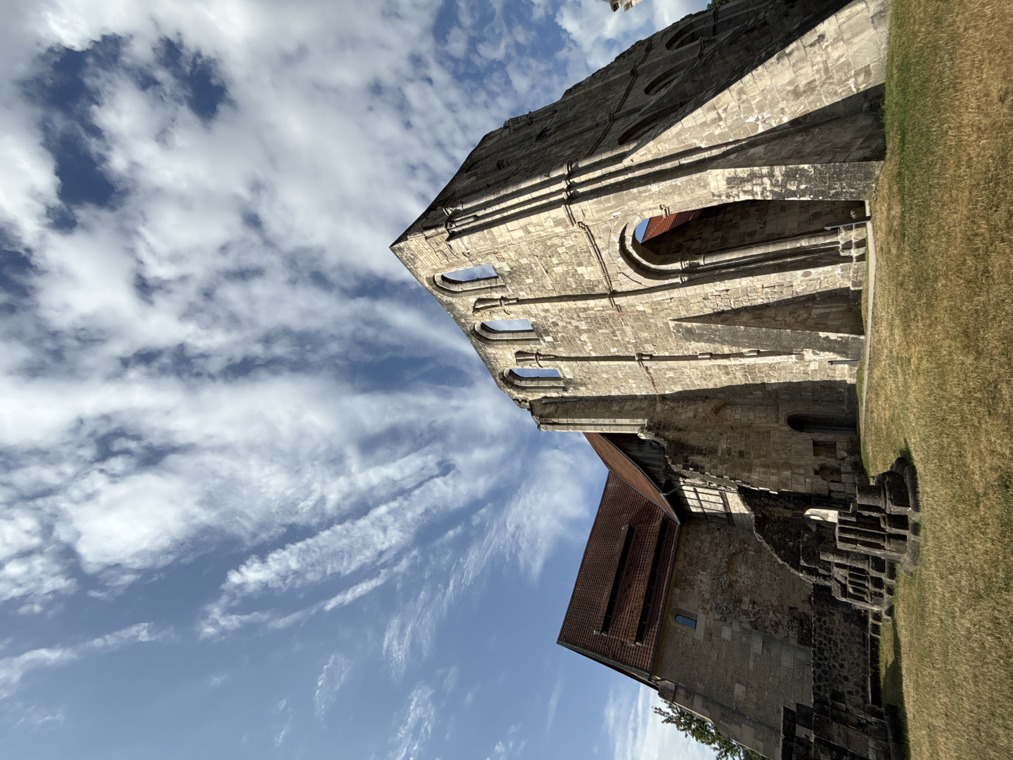 The image depicts a sunlit scene of ancient stone ruins against a backdrop of a bright blue sky dotted with wispy white clouds. The perspective is from below, looking upwards at the weathered stone structures, which appear to be part of a historical abbey or cathedral, suggested by the architectural style and the presence of arched windows and intricate stone carvings. The stone is a light grey and appears to be made of large, cut blocks.
In the foreground and to the right, a patch of dry, golden-brown grass suggests a sunny, possibly warm day. A portion of a building with a reddish-brown tiled roof is visible, juxtaposed against the older stone ruins. The overall setting evokes a sense of history and enduring architecture. The sunlight casts strong shadows, indicating it's likely midday or afternoon. There are no people visible in the image, focusing solely on the architectural remnants and the natural environment. The scene is serene and picturesque, highlighting the grandeur of the past against the timeless sky. The location is identified as Walkenried, Germany.