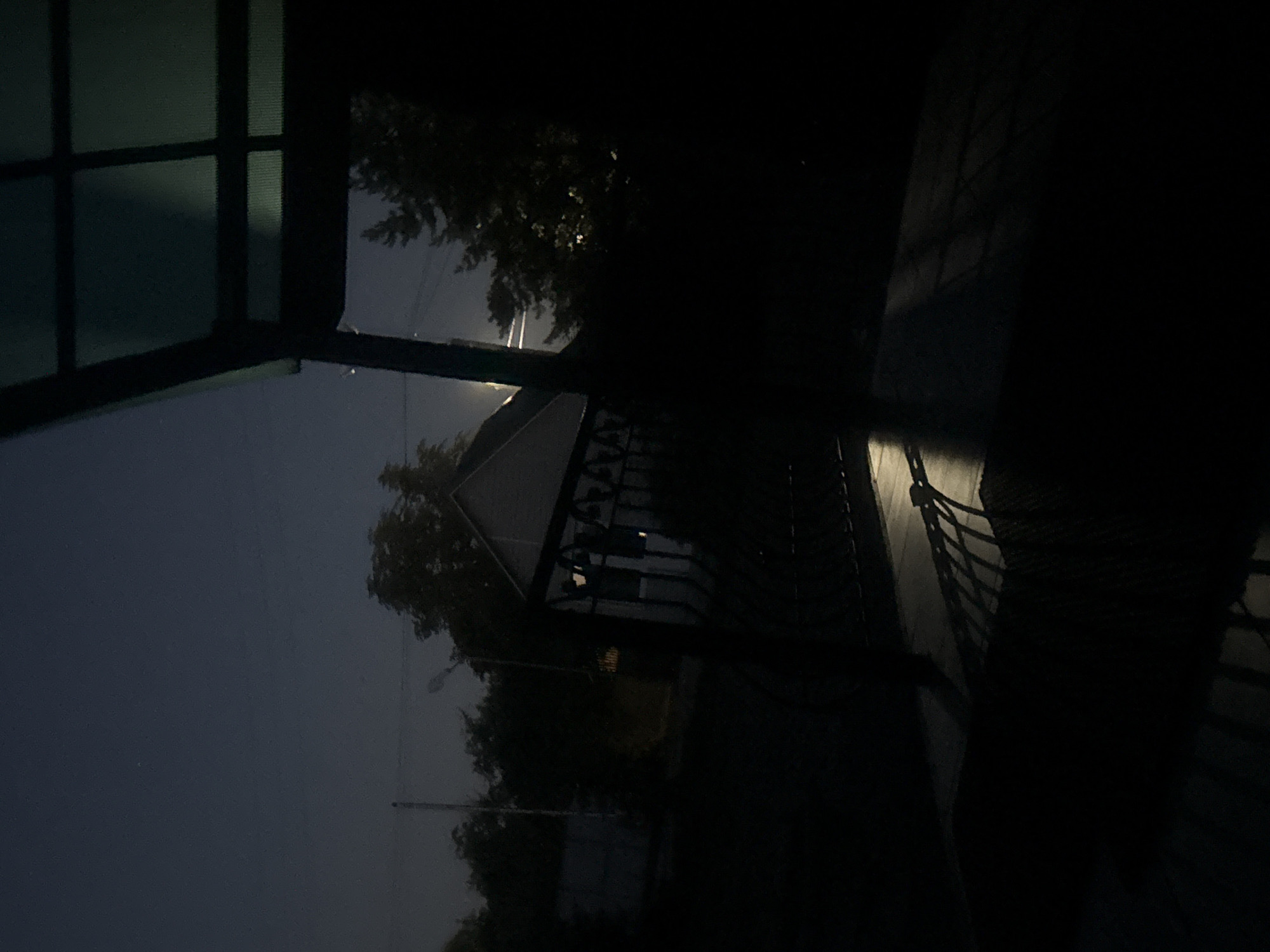 A night-time shot of a dark, outdoor scene, likely a backyard or patio area, shows a shadowy environment with limited illumination. In the foreground, a dark, possibly metal, fence or railing casts a long, geometric shadow across a paved surface. The paving stones are visible in the shadow, indicating a textured ground. Above and to the right, a portion of a dark green, possibly polycarbonate, roof structure is visible, with its grid-like internal supports casting faint shadows. Beyond the fence, a house with a light-colored gable roof is visible in the mid-ground, partially obscured by trees. The trees are dark silhouettes against a deep blue, twilight sky. A faint light source, possibly a streetlamp or distant light, illuminates a small area near the house and casts a glow through the trees. The overall atmosphere is somber and quiet.