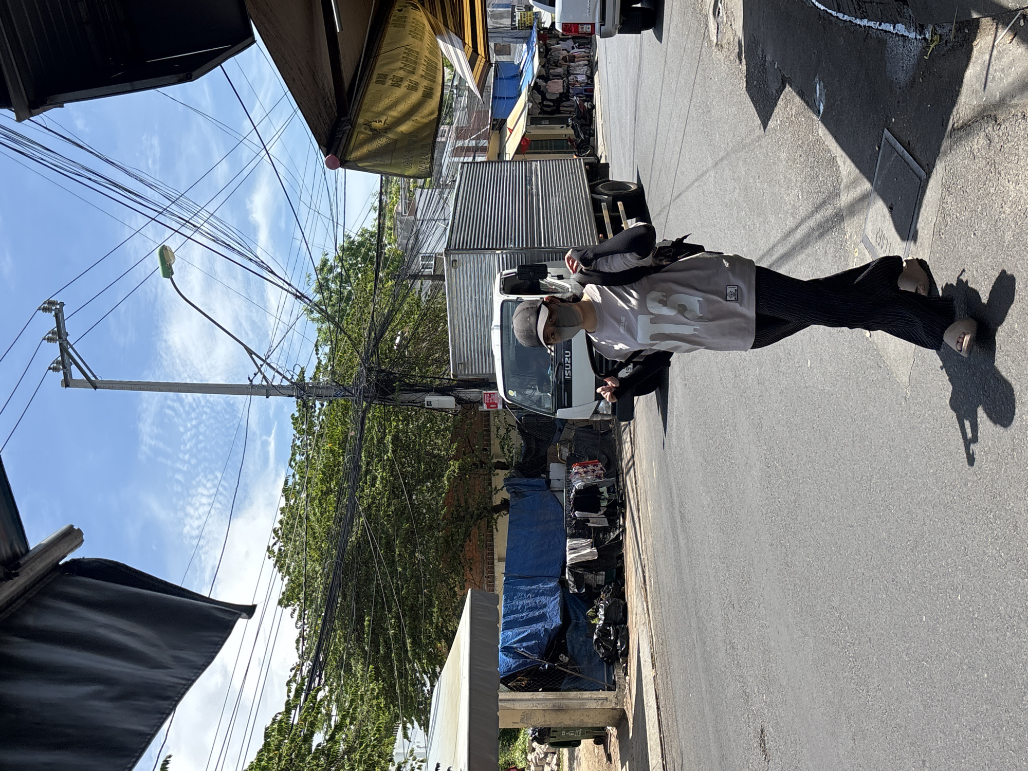 A medium shot captures a woman wearing a face mask, a grey t-shirt with "BTS" printed on it, dark pants, and sandals, walking on a street in Nha Trang, Vietnam. She is carrying a backpack and has a jacket draped over her shoulders. Behind her, an Isuzu truck is parked on the street. To the left of the truck, a makeshift stall is set up with blue tarps, clothing hanging on a rack, and black trash bags. The street is paved, and trees with dense foliage line the background. Overhead, a network of electrical wires and a street lamp are visible against a blue sky with scattered clouds. A yellow awning with text in Vietnamese is prominent on the right side of the frame. The scene appears to be a commercial area with shops and street vendors.