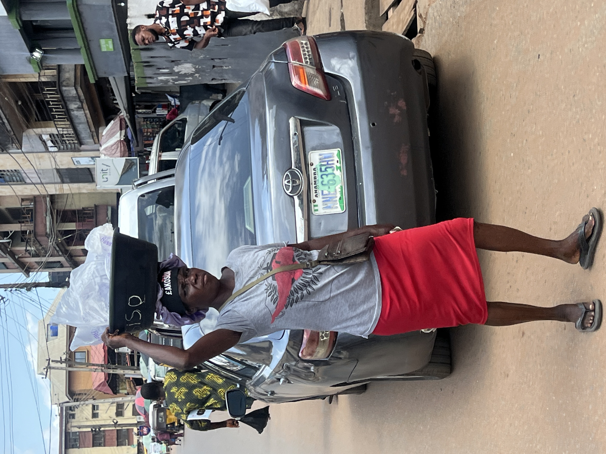A street scene in an unidentified city shows a woman carrying goods on her head, standing next to a parked gray Toyota car. The woman is wearing a gray t-shirt with a red design, a red skirt, and sandals. She also has a small purse slung across her shoulder. On her head, she carries a black tray loaded with plastic bags, and the word "ISO" is written in white on the side of the tray.
The car is parked on the side of the road. The license plate is visible and reads "ANAMBRA WNE-635HW". The area appears to be a bustling commercial district. Buildings are visible in the background, and power lines crisscross overhead. Other people are visible in the background, walking along the street.