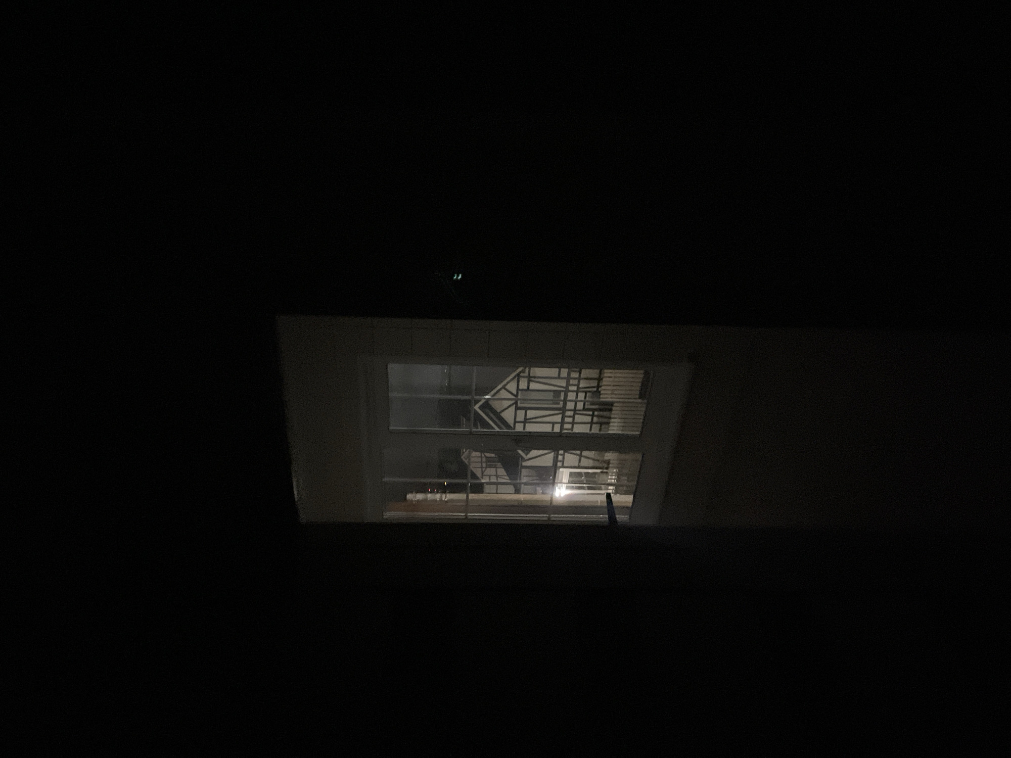 The image shows a night scene with a view through a window. Outside the window, there is a structure that appears to be scaffolding or some type of metal framework. Inside, the room is dark, but the light from outside illuminates the window frame. Based on the location provided, this scene is likely in Хлябово, Russia.