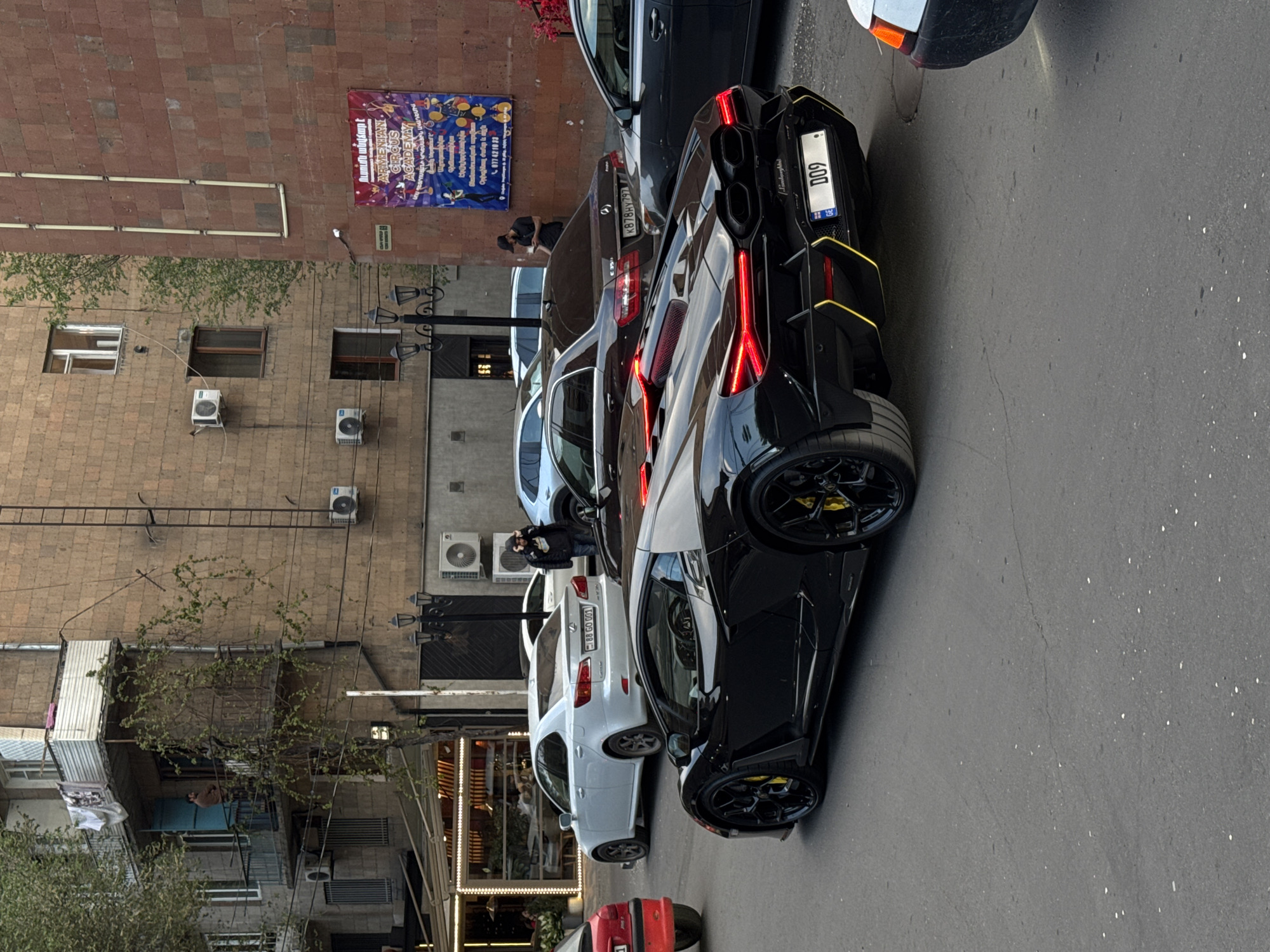 This image captures a vibrant street scene in an urban environment, likely in Yerevan, Armenia, as indicated by the location context and visible text. The time of day appears to be late afternoon or early evening, with soft lighting and some shadows present.
The primary subjects are several parked cars and surrounding buildings. In the foreground, a sleek black Lamborghini with yellow accents is prominently featured, displaying a license plate "D09" with a small Armenian flag emblem. Next to it, a dark-colored Mercedes-Benz C-Class sedan is visible, bearing the license plate "K 878 HY 797". Further down the street, a white Lexus sedan with license plate "88 GO 001" (also with an Armenian flag emblem) is parked. Other vehicles are partially visible, including a red car at the very bottom left and another car behind the Mercedes.
Several people are scattered throughout the scene. One individual in a black jacket and hat is seen near the Mercedes, possibly observing the cars. Another person is standing by the white Lexus.
The background consists of multi-story brick and concrete buildings. These structures feature numerous windows, some with air conditioning units attached to their exteriors, and a few small balconies. Green foliage, possibly from trees or vines, can be seen along some building walls and balconies. Street lights with ornate designs are also visible.
A notable detail is a large colorful banner affixed to one of the brick buildings. The text on the banner, which is in Armenian and English, reads "Ուսումն անվճար է" (Education is free), "ARMENIAN CIRCUS ACADEMY", "Օրիգինալ ժանր և այլն" (Original genre etc.), and a phone number "077 42 10 33". This banner, along with the distinct Armenian license plates, strongly anchors the location. The overall atmosphere suggests a typical city street, with no specific event other than the daily flow of urban life.