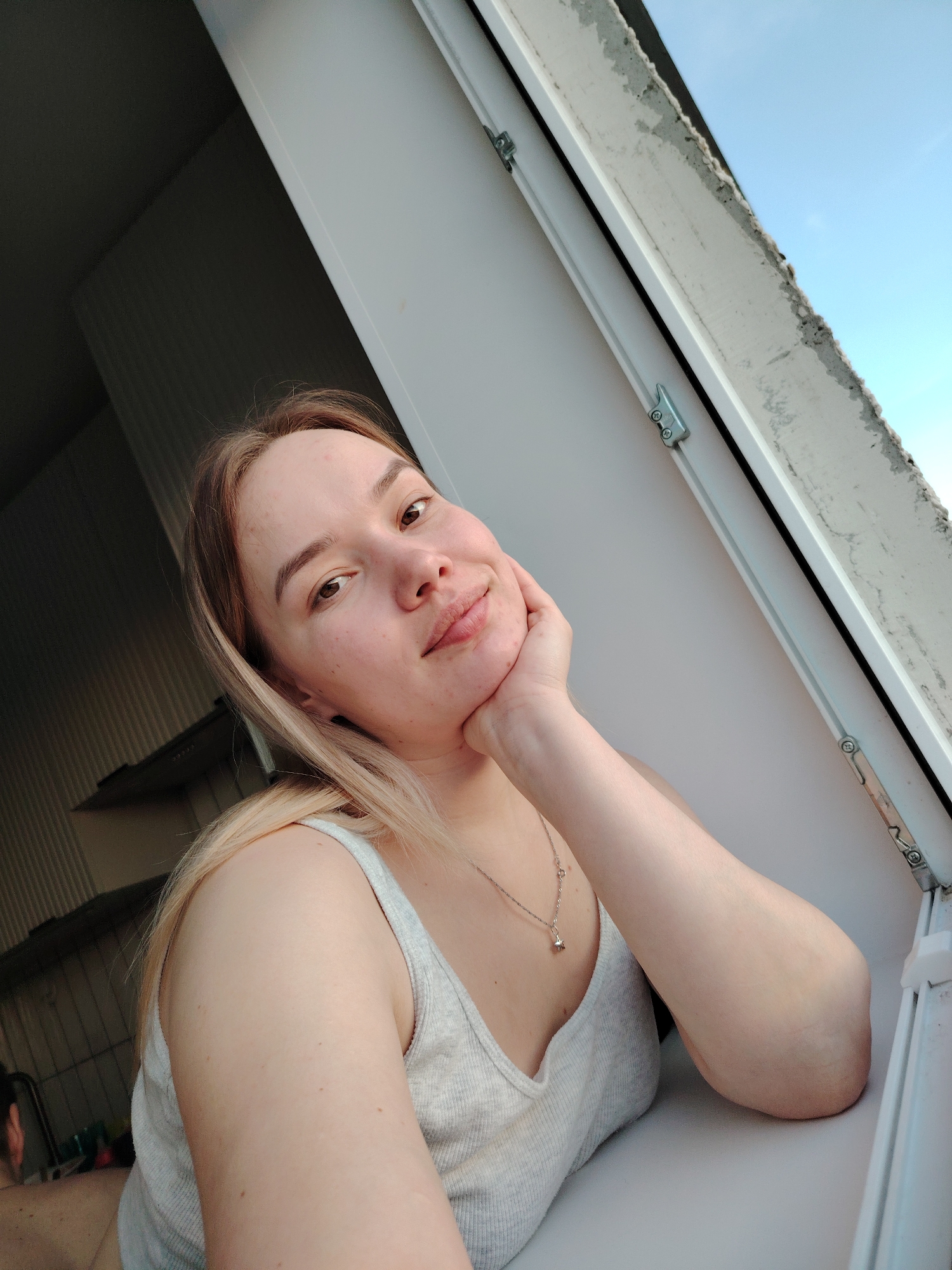 A woman with blonde hair and a grey tank top is leaning on a window sill, looking out a window. She rests her chin on her hand. The window frame is white, and a small portion of a blue sky is visible through the window. The background is partially obscured, but appears to be an interior space with shelves. The image may have been taken in Марковский, Russia.