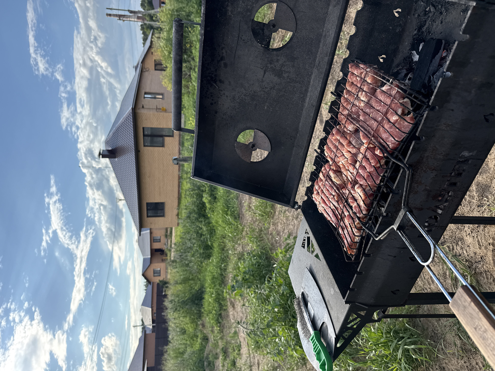 A close-up, eye-level outdoor shot captures a black metal barbecue grill filled with rows of raw meat skewers. The skewers are arranged in parallel lines across a metal grate, with the meat appearing pink and unfettered by seasoning. The grill itself is situated in what appears to be a rural or suburban setting, with a patch of dry earth and green grass visible in the foreground. In the background, several single-story houses with brick exteriors and tiled roofs are visible, nestled amongst lush green foliage. A bright blue sky with scattered white clouds completes the scene, suggesting a pleasant, possibly summer, day. The barbecue tools, including a grill brush and scraper with a green handle, rest on the side of the grill. The general ambiance is one of preparing for an outdoor meal. The location is identified as Chaikovsky, Russia.