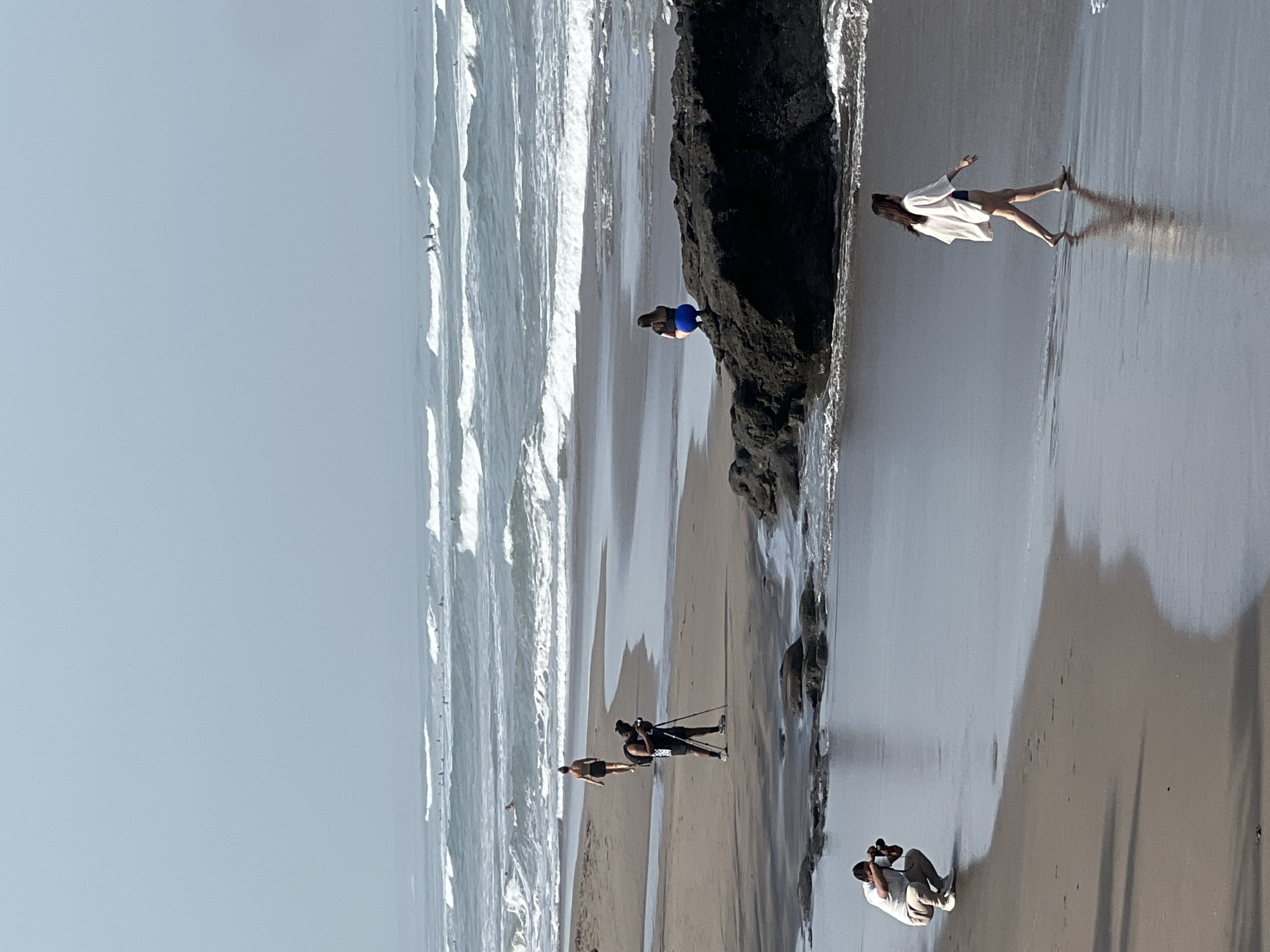 This is an outdoor, eye-level, medium shot of a beach with several people and the ocean in the background. The scene is the city of North Kuta, Indonesia. The water is calm with small waves. Several surfers are visible in the distance. On the shore, a person is walking in the shallow water on the left side of the frame. To the right of the frame, a person is sitting on a dark rock formation. Further in the background, a person is standing on the beach with a camera on a tripod. In the foreground, a person is crouching and taking a photograph. The beach is a mix of sand and some dark, wet patches.