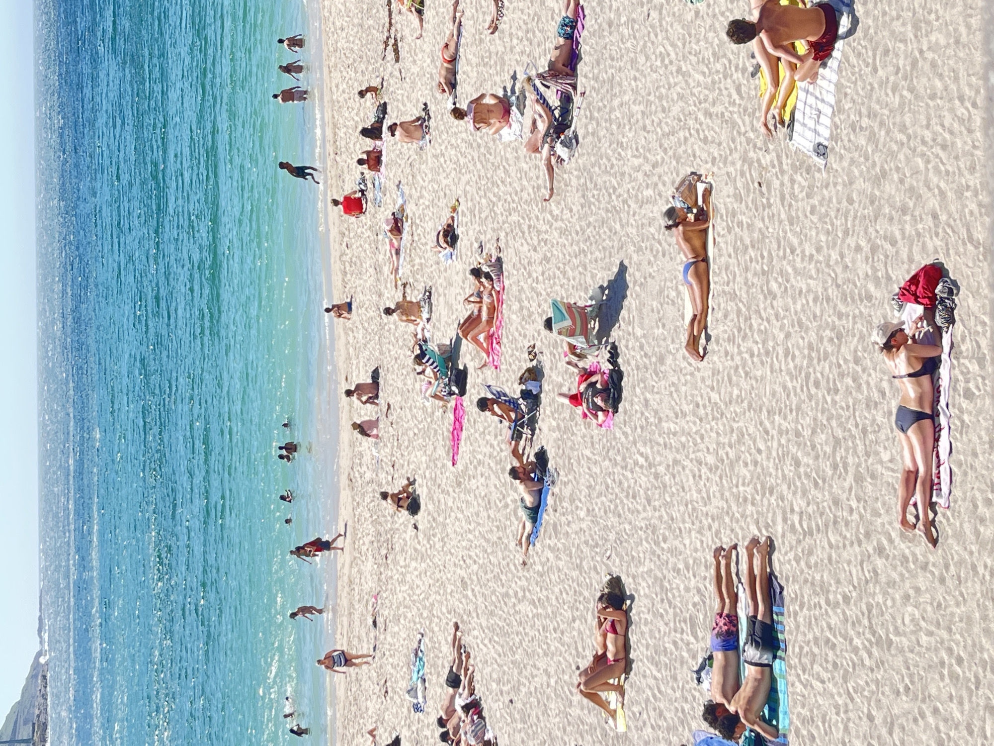 This is an aerial, high-angle shot of a crowded beach on a sunny day. The turquoise ocean on the left contrasts with the warm, sandy beach dotted with people. The sun appears high in the sky, casting short shadows, indicating it's likely midday. The beach is filled with individuals and small groups sunbathing on towels, swimming in the shallow water, or walking along the shoreline.
The subjects are primarily people in swimwear, relaxing and enjoying leisure activities. There are various beach objects scattered across the sand, including towels of different colors and patterns, a beach umbrella, and bags. The scene depicts a typical summer day at a popular beach destination.
Notable details include the clear, bright sunlight illuminating the scene and the calm, vibrant color of the sea. The density of people suggests a popular spot, likely during peak tourist season. The overall mood is one of relaxation and enjoyment. No visible text is present in the image. The context provided places this scene in A Coruña, Spain.