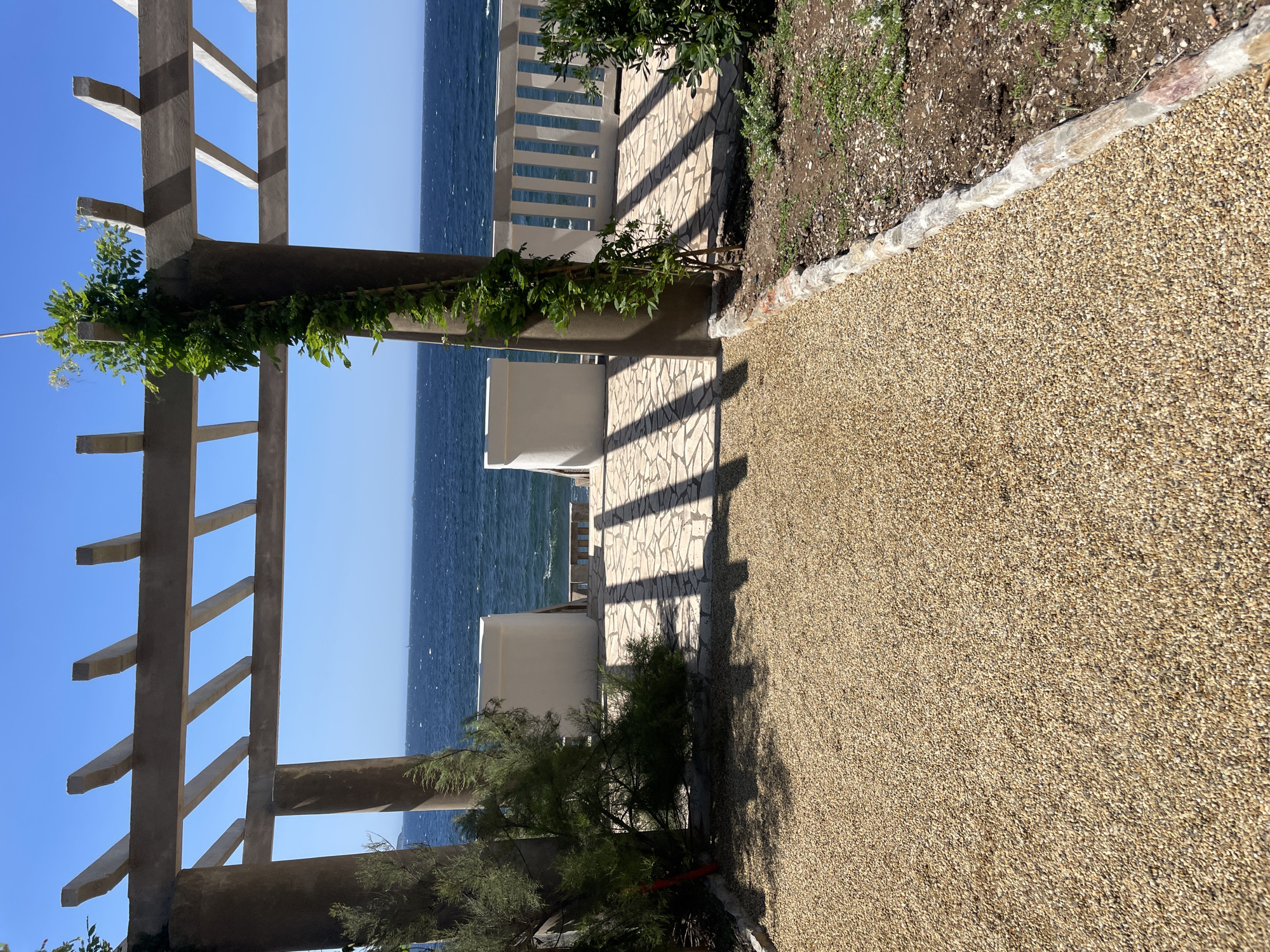 This is a photograph taken outdoors in Carqueiranne, France. The image shows a view through a pergola structure with concrete pillars and beams. The pergola frames a clear blue sky and a body of water, likely the sea, which appears choppy. In the foreground, a path covered in small, light-colored gravel leads towards the water. To the sides of the path are some low-lying green shrubs and a larger, sparse bush on the left. The paved area beyond the gravel path is made of irregular stone slabs, casting distinct shadows from the pergola. There are white, low walls on either side of a set of steps leading down to a white railing overlooking the water. A vine with green leaves is growing up one of the concrete pillars on the right. The overall scene depicts a sunny day with a serene, coastal atmosphere.