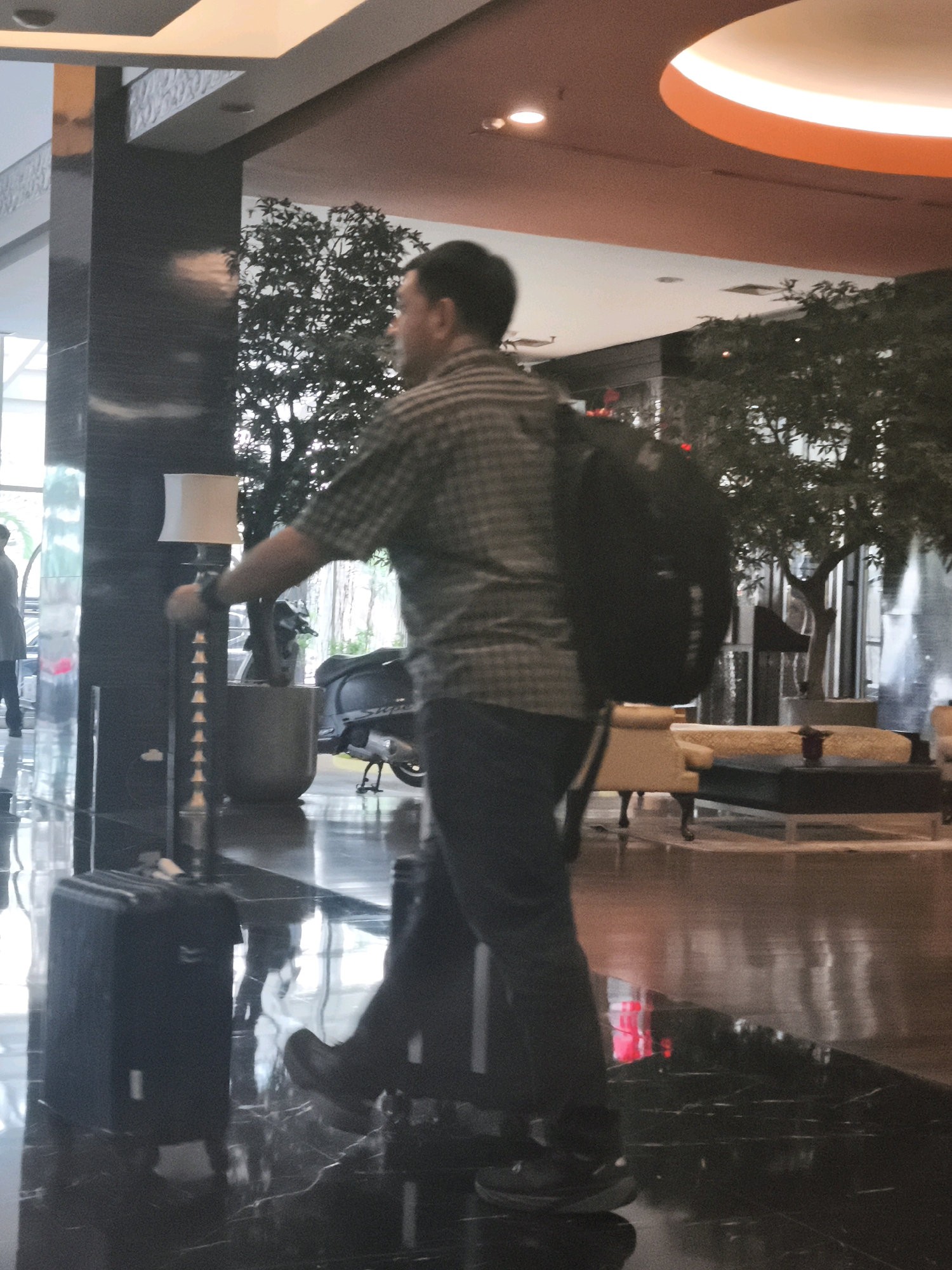 A man wearing a checkered shirt and dark pants walks through a hotel lobby, pulling a black suitcase behind him with his left hand. He is also carrying a black backpack. In the background, a motorcycle is parked near potted plants. The lobby features a polished black floor, a tall floor lamp with a white shade, and seating areas with yellow upholstered furniture. The overall lighting suggests an indoor commercial space. Based on visual cues and typical architectural styles in Palembang, Indonesia, this scene is likely occurring in a hotel lobby in the city.