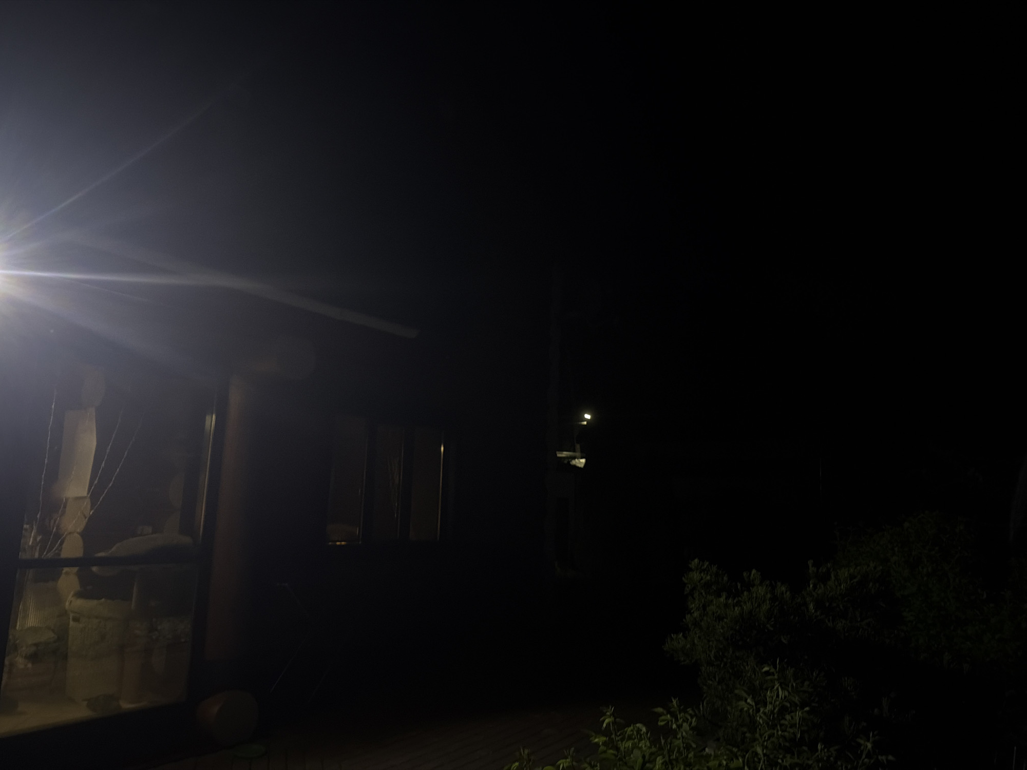 The photograph, taken at night, depicts a building with a partially visible facade due to a bright light source to the left. A window is seen, behind which are visible light-colored objects. To the right of the building are dark trees and shrubbery. A pole with a light fixture is observed in the middle-ground. The overall scene is dimly lit. The location is given as Chaikovsky, Russia.