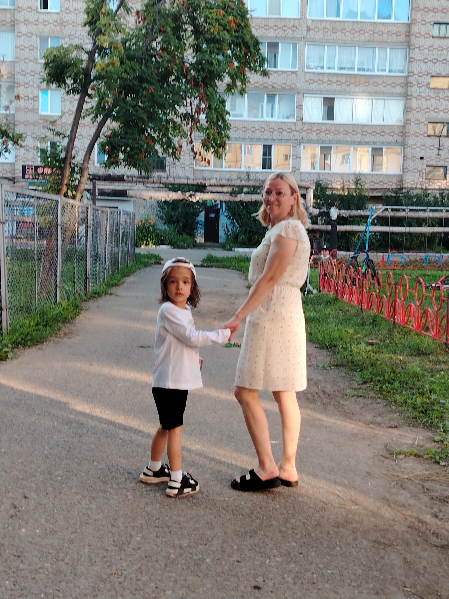 An outdoor, eye-level, full shot depicts a woman and a young child holding hands and standing on a paved pathway. The woman, positioned on the right, wears glasses and a light-colored, short-sleeved dress with a subtle pattern. The child, on the left, has dark hair and is wearing a white long-sleeved shirt, black shorts, a white cap, and black and white sneakers. Behind them, a multi-story apartment building with light-colored brickwork and numerous windows dominates the background. A tree with green leaves and reddish berries stands in front of the building. A metal fence runs along the left side of the pathway, and on the right, a red circular-patterned fence separates the pathway from a grassy area with some playground equipment. The scene appears to be located in the city of Yanaul, Russia.