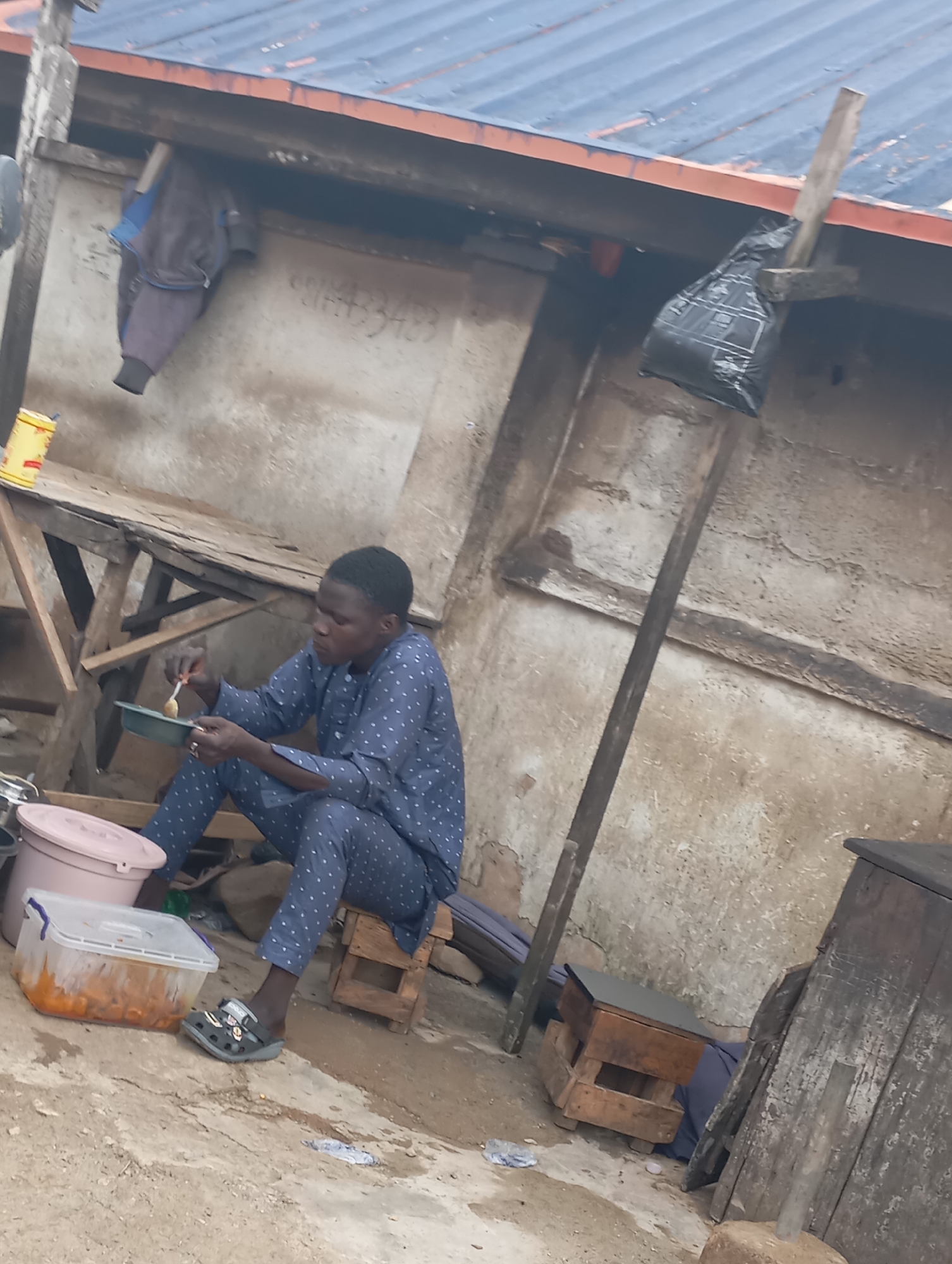 In Jos, Nigeria, a young man in a blue dotted outfit is seated on a small wooden stool outdoors, eating from a green bowl. He holds a spoon in his right hand, bringing food to his mouth. To his left, a pink plastic basin and a transparent plastic container filled with food are visible on the ground. Behind him, a rickety wooden structure with a corrugated iron roof suggests a rustic setting, possibly a makeshift stall or dwelling. A grey jacket is hung on a wooden beam above a roughly plastered wall, which shows signs of wear and tear. A black plastic bag is secured to a wooden pole near the roofline. The ground is uneven concrete, with scattered debris. The overall scene depicts a simple, everyday moment in an outdoor environment.