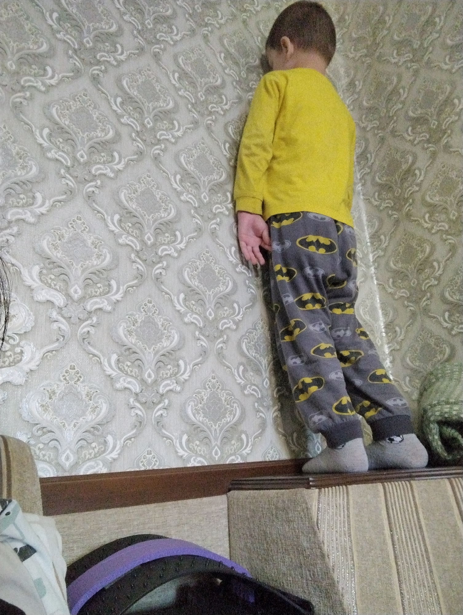 The image shows a young boy standing with his back to the camera, leaning against a wall with ornate, damask-patterned wallpaper. He is wearing a yellow long-sleeved shirt and gray pajama pants featuring a repeated pattern of Batman logos in yellow and black. He has on light gray socks. The boy's left hand is visible, resting on his leg.
The boy is standing on the top edge of a piece of furniture, possibly a sofa or a bench, which is upholstered in a striped fabric. In the foreground, a purple and black strap or handle is visible, suggesting a bag or some other object. To the right of the boy, a green knitted item, likely a hat or scarf, is partially visible. The lighting appears to be indoor, with no strong indications of time of day or weather conditions. The wallpaper and the furniture suggest a domestic setting. The overall mood of the image is quiet and perhaps a little somber due to the boy's posture and the muted colors. There is no visible text in the image. The location context is Kingisepp, Russia.