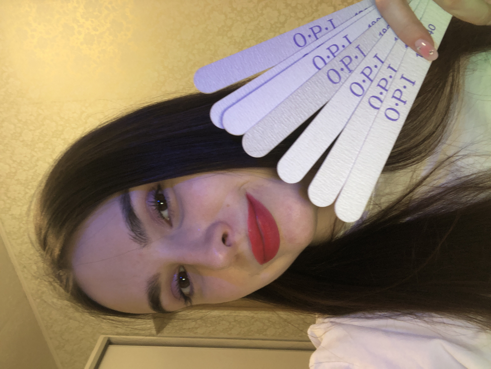 A young woman with long, dark hair is depicted in an indoor setting, looking directly at the viewer with a gentle smile. Her head is tilted to the side, revealing her dark eyes, pronounced eyebrows, and red lipstick. She is wearing a light-colored top.
In her right hand, she holds a fan of several light purple and white O.P.I brand nail files. The "O.P.I" logo is prominently displayed in purple text on each file. Visible grit numbers on the files include "100" and "240". Her right index fingernail features a decorative manicure with a pinkish base and small, reflective embellishments.
The background consists of a warm yellow wall adorned with a subtle floral or textured pattern. The lighting appears warm and artificial. The overall scene suggests a context related to nail care or beauty. This image captures a moment in Noviy, Russia.