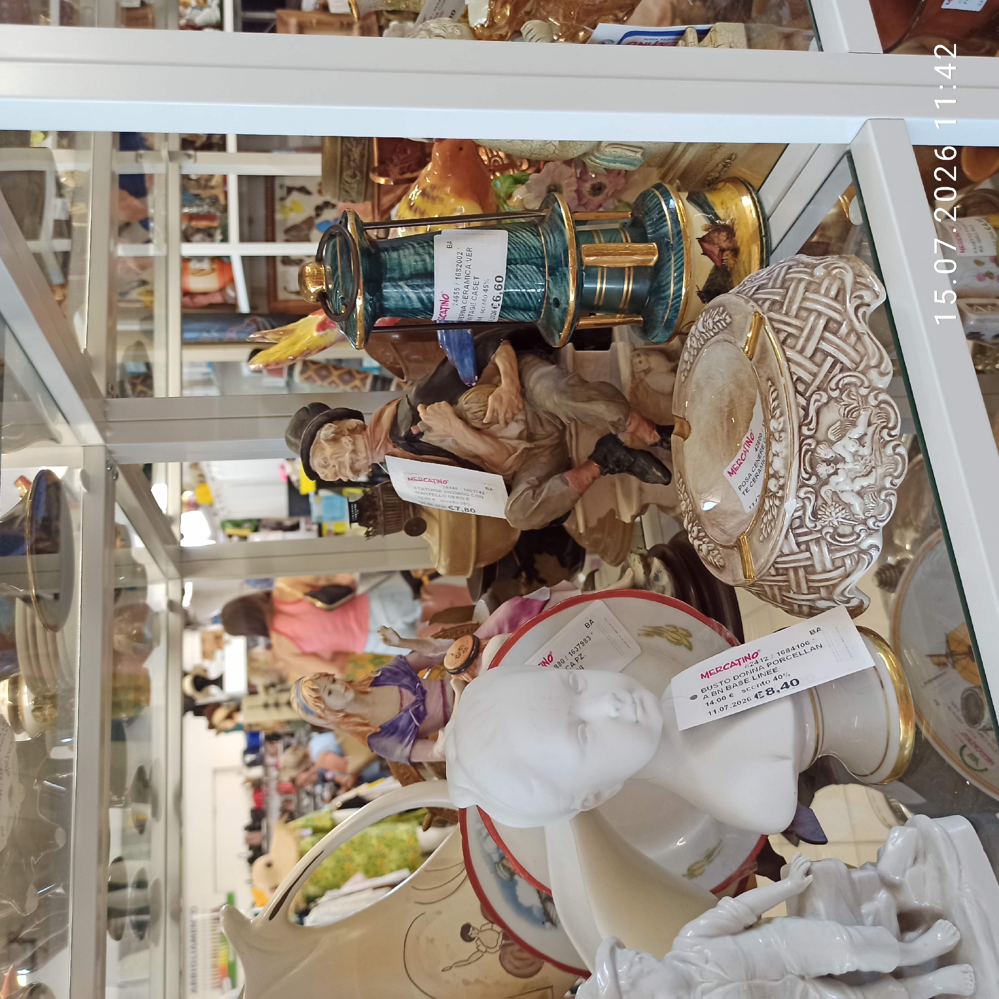 The image depicts a display of antique or decorative items in a shop, possibly a vintage store or flea market. The items include ceramic figurines, decorative plates, an ashtray, and a lamp. In the foreground, a white ceramic bust of a young girl with a golden rimmed base is visible, with a price tag indicating "MERCATINO" and a price of €8.40. To the left of the bust is a white ceramic figurine of a young boy. Behind these, a woman in a purple dress is seated, holding a tambourine, with a price tag on her base as well. To the right of the bust is an ornate, woven-patterned ceramic ashtray with depictions of cherubs. Further to the right is a teal and gold ceramic oil lamp with a price tag showing "MERCATINO" and "€6.60". In the background, other assorted decorative items are displayed on shelves, and several people are browsing in the store. One woman in a pink top is visible in the mid-ground. The overall environment suggests an indoor retail space with glass shelving. The date stamp at the bottom right corner indicates "15.07.2026 11:42". There is no explicit indication of the location being Molfetta, Italy, within the image itself.