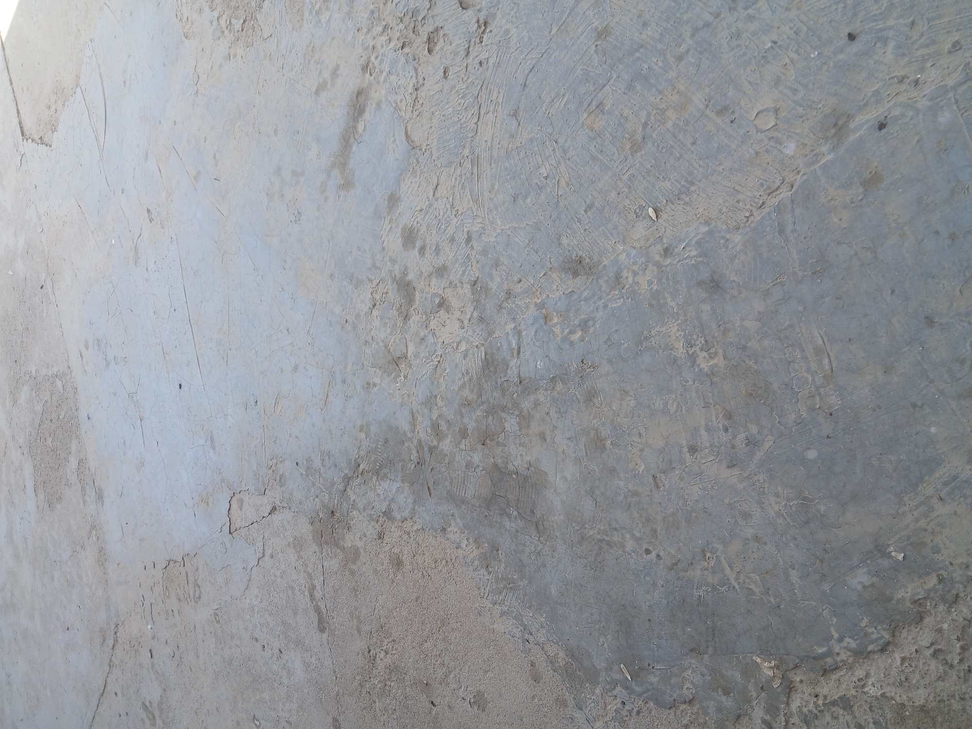The image displays a close-up, overhead view of a cracked concrete surface. The concrete is weathered and appears to be an outdoor area, possibly a pathway or a yard. Patches of sand and dirt are visible on the surface, particularly on the left side of the image. There are numerous fine cracks and some larger fissures running across the concrete, indicating wear and tear. The lighting suggests it is daytime, casting a slight shadow and highlighting the texture of the material. The environment is characteristic of an unmaintained or utilitarian outdoor space. No people or specific objects are discernible in the image. The location is the rural district of Kyzylzhar, Kazakhstan.