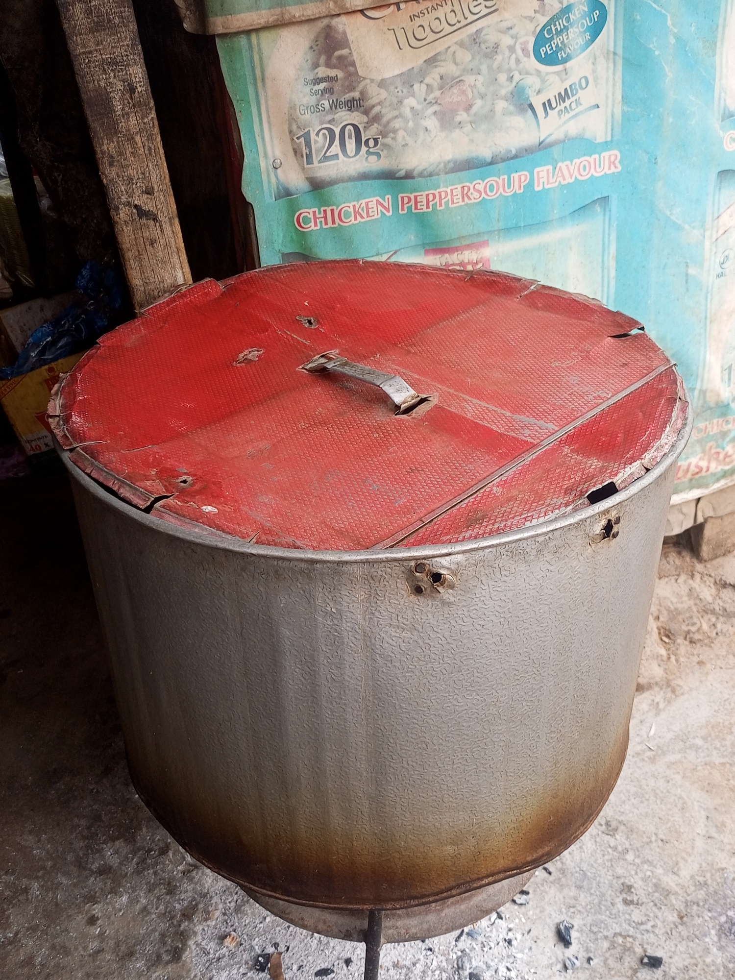 An eye-level medium shot reveals a large, cylindrical, metal container with a red, damaged lid in an outdoor environment, likely in Maiburiji, Nigeria, given the surrounding context. The container, resting on a metal stand, shows signs of wear, with a dark stain encircling its base. The red lid is also damaged, with tears and holes. In the background, noodle packaging is visible, including a packet labelled "Chicken Peppersoup Flavour". To the left of the container is a wooden post and some refuse. The ground is concrete, with scattered debris and remnants of what appears to be burnt material. The overall scene has an unkempt, utilitarian feel.