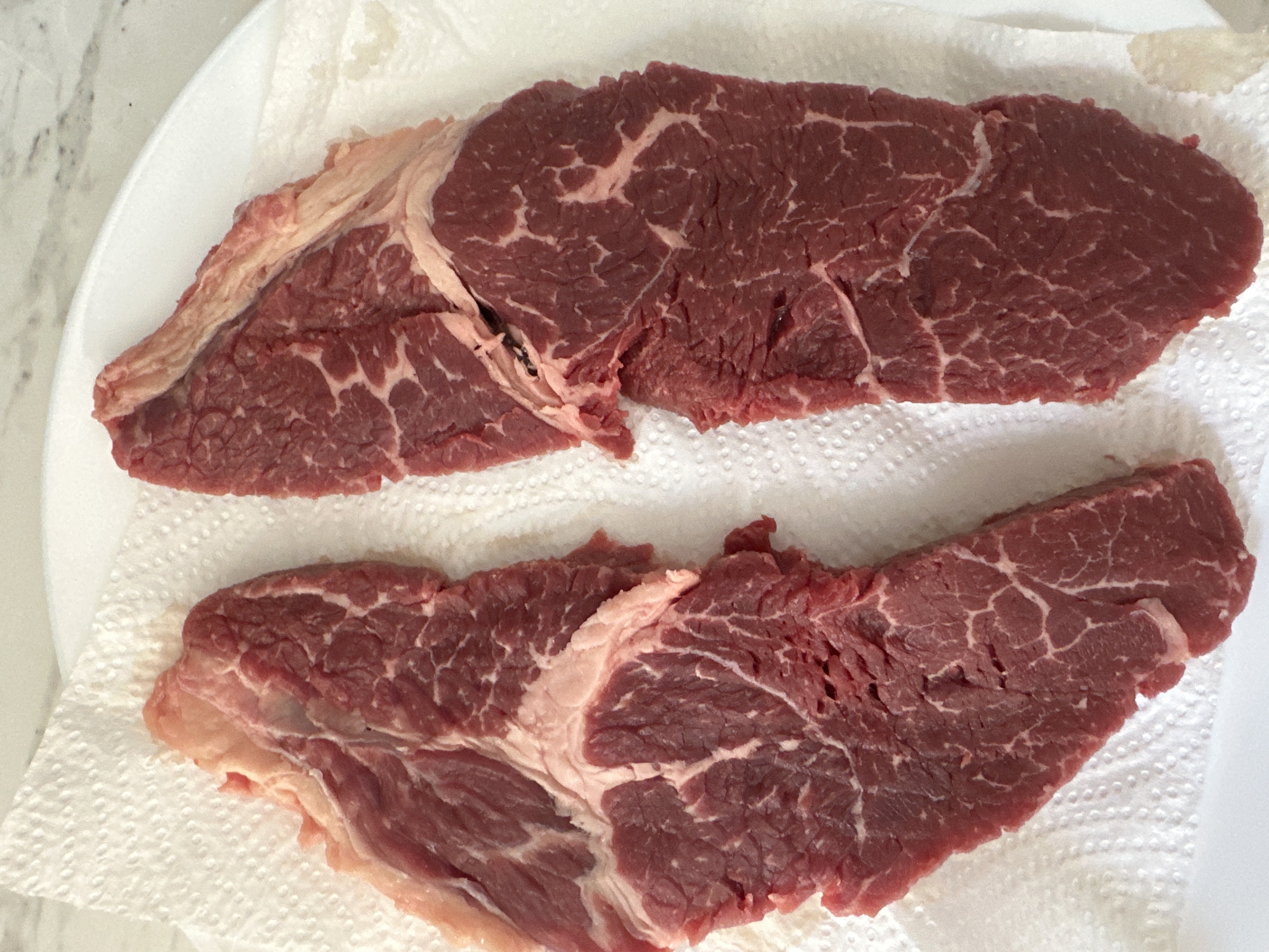 Two raw steaks are laid out on a white paper towel, which is on top of a white plate. The steaks are a deep red color with visible marbling. The steak on the left is oriented vertically, with the top of the steak pointing upwards. It has a lighter colored strip of fat along its left edge. The steak on the right is also oriented vertically, to the right of the first steak. It also has a strip of fat along its left edge. The background is a light-colored, speckled surface, possibly a countertop. The image does not provide any information about the city of Moortown, Ireland.