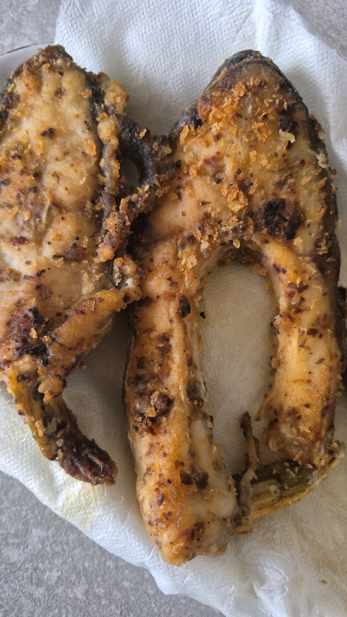 Two fried fish steaks are presented on a white paper towel. The steaks are golden brown with visible seasoning and some darker, charred bits. One steak is cut in a U-shape, with the bone exposed in the center. The other steak is a more irregular, curved piece. The fish appears to be resting on a light gray, textured surface, possibly a countertop or table, with a white paper towel underneath. The scene is a close-up, overhead shot. This image does not depict any specific location in Krauja, Latvia.