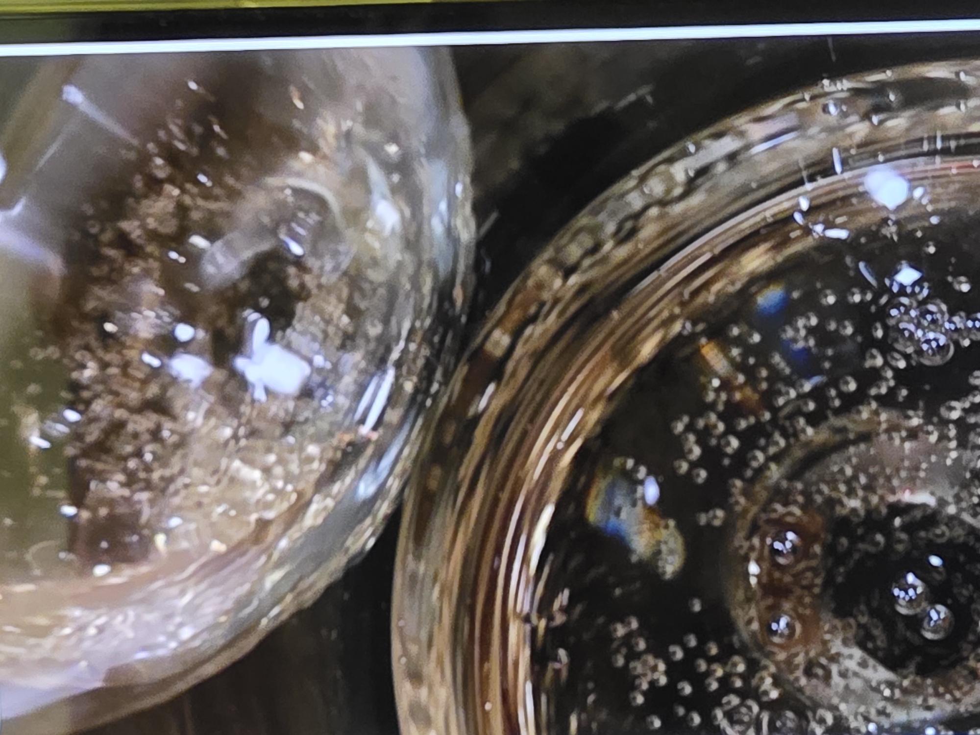 The image is a close-up, top-down view of two glasses filled with a bubbly liquid, possibly champagne or sparkling wine. One glass is positioned above and behind the other. The liquid in both glasses appears light in color with small bubbles rising to the surface. The glass in the foreground has more visible bubbles, and a faint rainbow-like refraction is visible on its surface. The background is dark and out of focus. The scene is indoors, with the lighting suggesting a dimly lit environment. There are no people visible in the image.