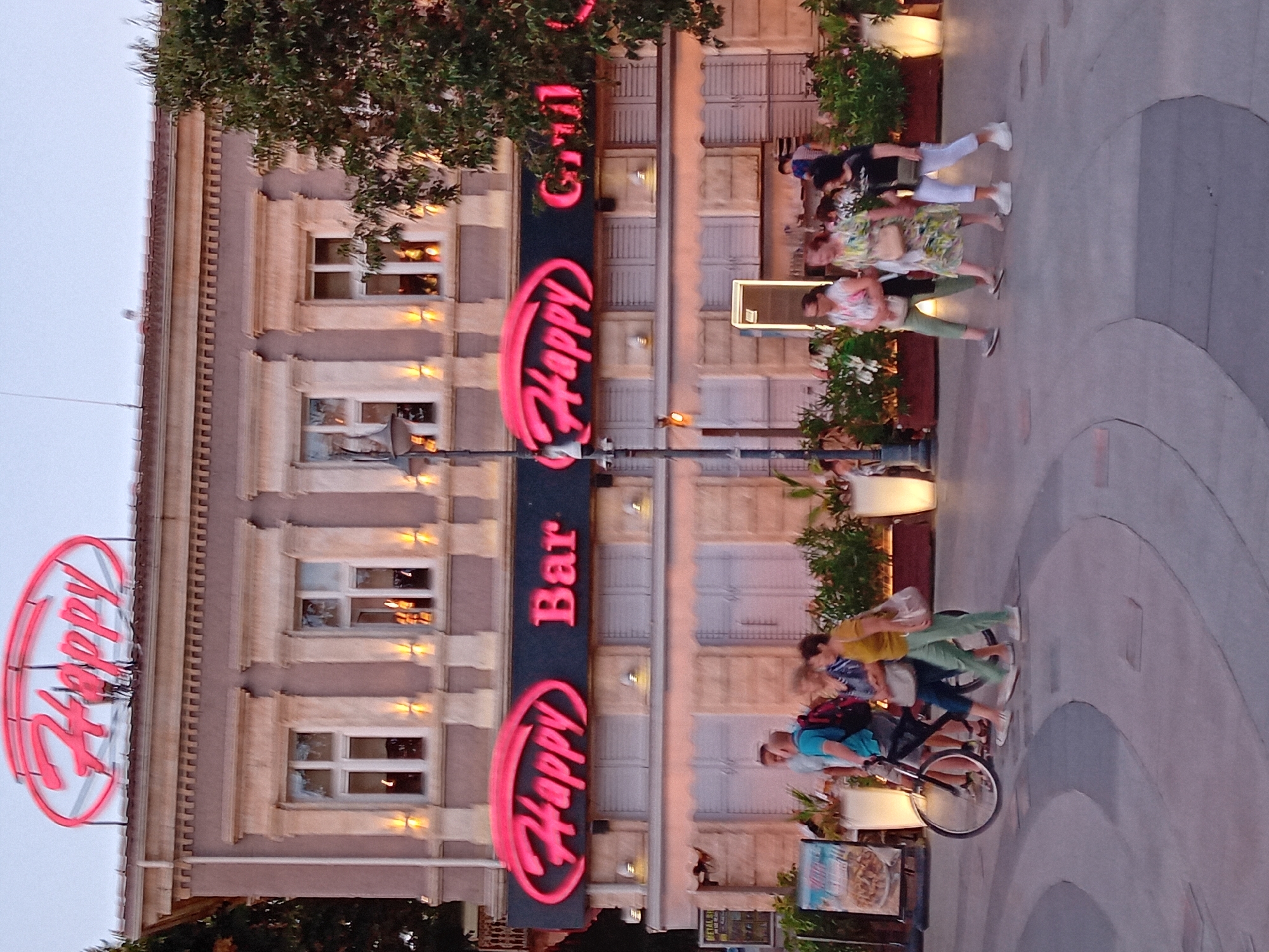 The image shows the exterior of "Happy Bar & Grill" in Varna, Bulgaria, at dusk. A large neon sign with "Happy" in pink letters is visible on the roof of the building, as well as a similar sign above the entrance that reads "Happy Bar Grill". The building has a classical architectural style with columns and ornate window frames. In front of the building, several people are walking on a paved walkway. A man is pushing a bicycle, and three women are walking together. Green plants in planters are placed along the building's facade. The lighting suggests it is either early evening or late afternoon.