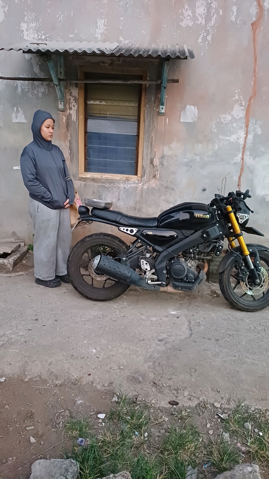 A full-shot, medium-angle, outdoor photograph shows a young woman standing next to a black Yamaha motorcycle parked on a cracked concrete surface. The woman, wearing a gray hooded jacket and light gray sweatpants, stands with her hands in her pockets, facing slightly away from the camera. The motorcycle, a sportbike, is parked on the right side of the frame. It has a black fuel tank with the "YAMAHA" logo in gold lettering, black body panels, yellow front suspension forks, and a black exhaust pipe. Behind the woman and motorcycle is a distressed, light-colored building with a corrugated metal awning above a window with horizontal blue panes. The environment appears to be a gritty urban setting in Cibinong, Indonesia, with patches of sparse grass growing through the concrete in the foreground. The lighting suggests it is daytime, possibly late afternoon.