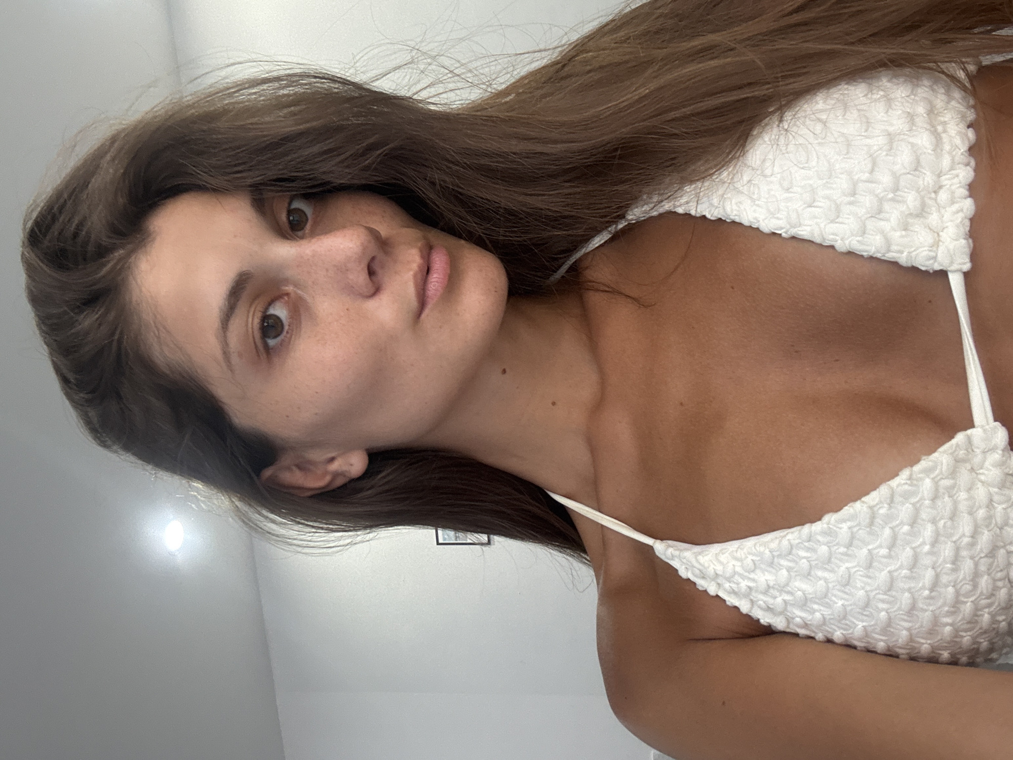 A woman with long brown hair poses in a white textured bikini top. She has fair skin with freckles and is looking directly at the camera. The background appears to be a plain white wall, likely indoors. The picture is slightly rotated counterclockwise. The location in the city of Đà Nẵng, Vietnam, is not possible to determine from the image.