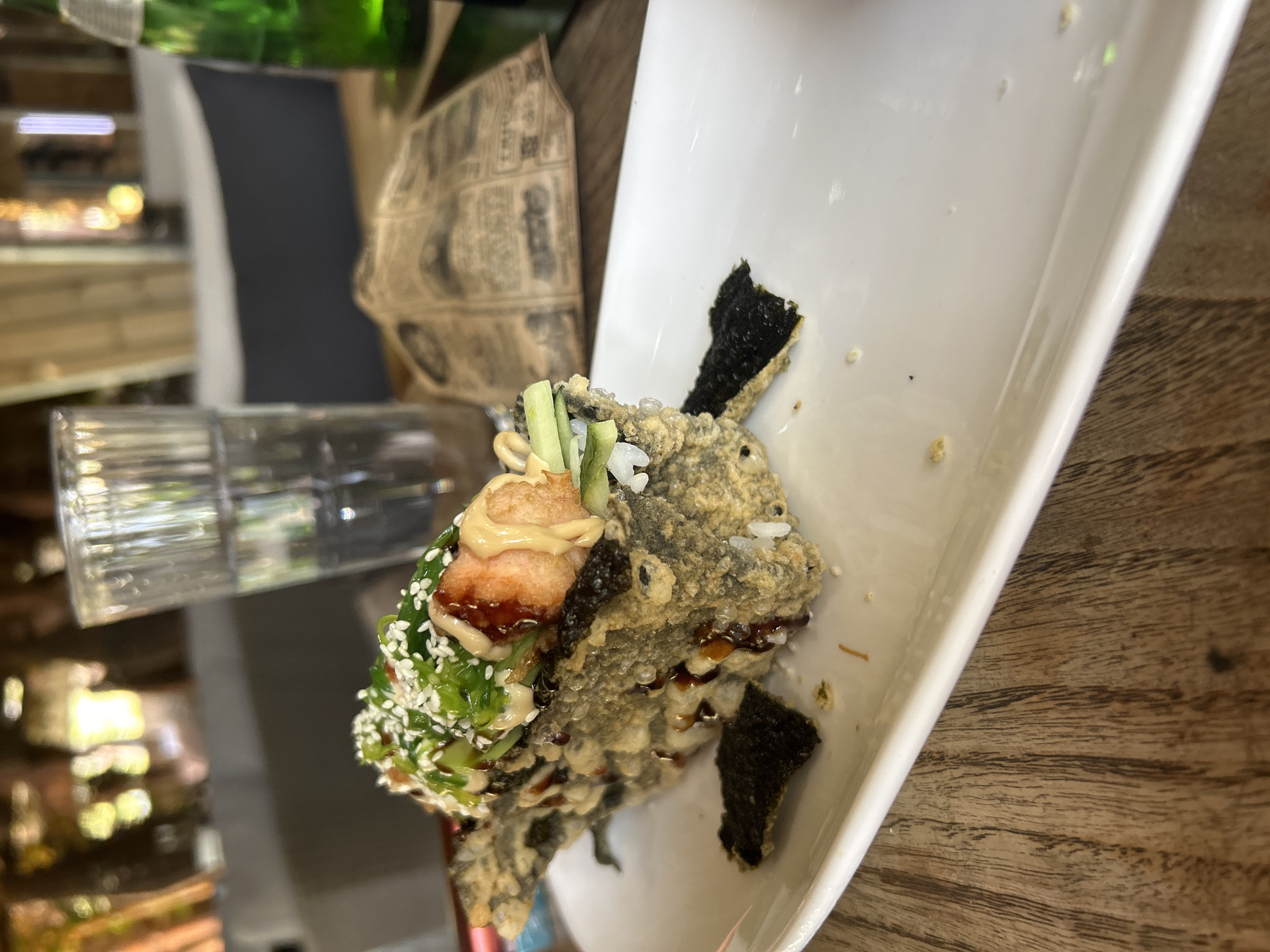 A single, large, deep-fried sushi roll is presented on a white rectangular plate, which is placed on a wooden table. The sushi roll has a crispy, golden-brown exterior, likely tempura-battered, with visible pieces of nori seaweed. It is topped with what appears to be a piece of shrimp, green onions, sesame seeds, a drizzle of creamy sauce, and possibly some cucumber. There is also a dark sauce drizzled over the roll and the plate. In the background, a glass of water is visible, along with a green bottle and some crumpled paper with text. The overall environment suggests an outdoor dining setting, possibly a restaurant with a relaxed atmosphere, given the wooden table and the natural light filtering in. There is no indication of the city of Sofia, Bulgaria.