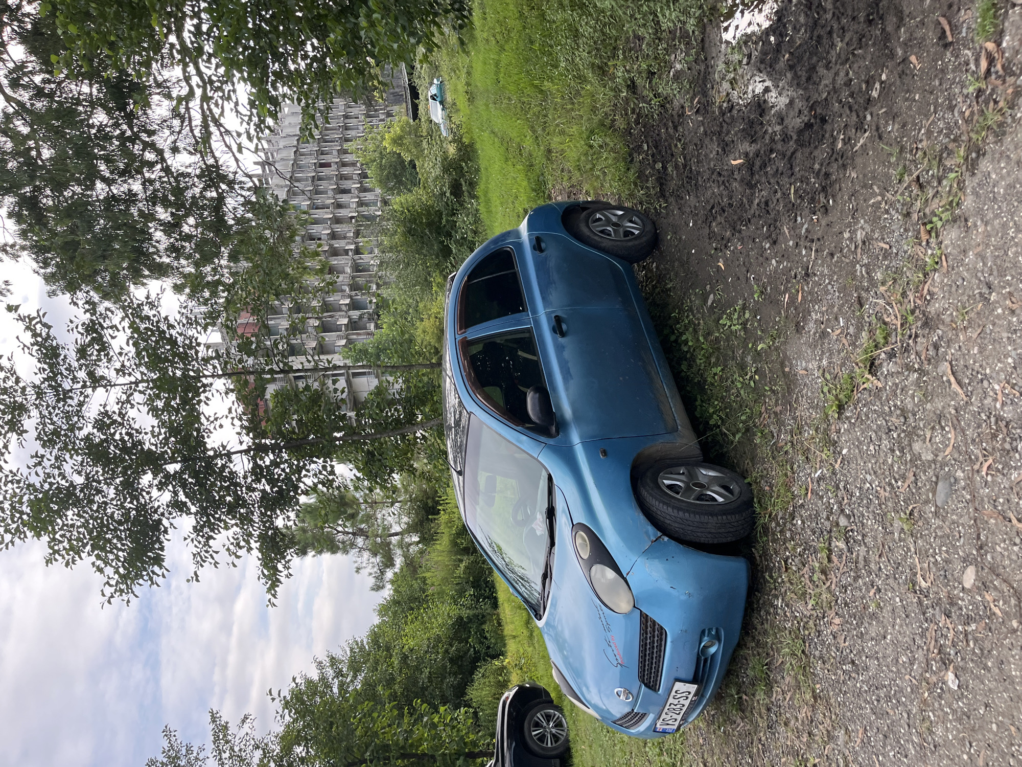 In Ureki, Georgia, a blue Nissan Micra, license plate VS-283-SS, is parked on a dirt and grass area next to a muddy patch. The car is angled towards the viewer's left. Behind the car, a multi-story abandoned building with many balconies is visible, surrounded by lush green trees and vegetation. The sky is partly cloudy. Another car, possibly green, is parked further in the background. A black SUV is partially visible on the far left of the frame. The overall scene suggests a somewhat overgrown and neglected urban environment.