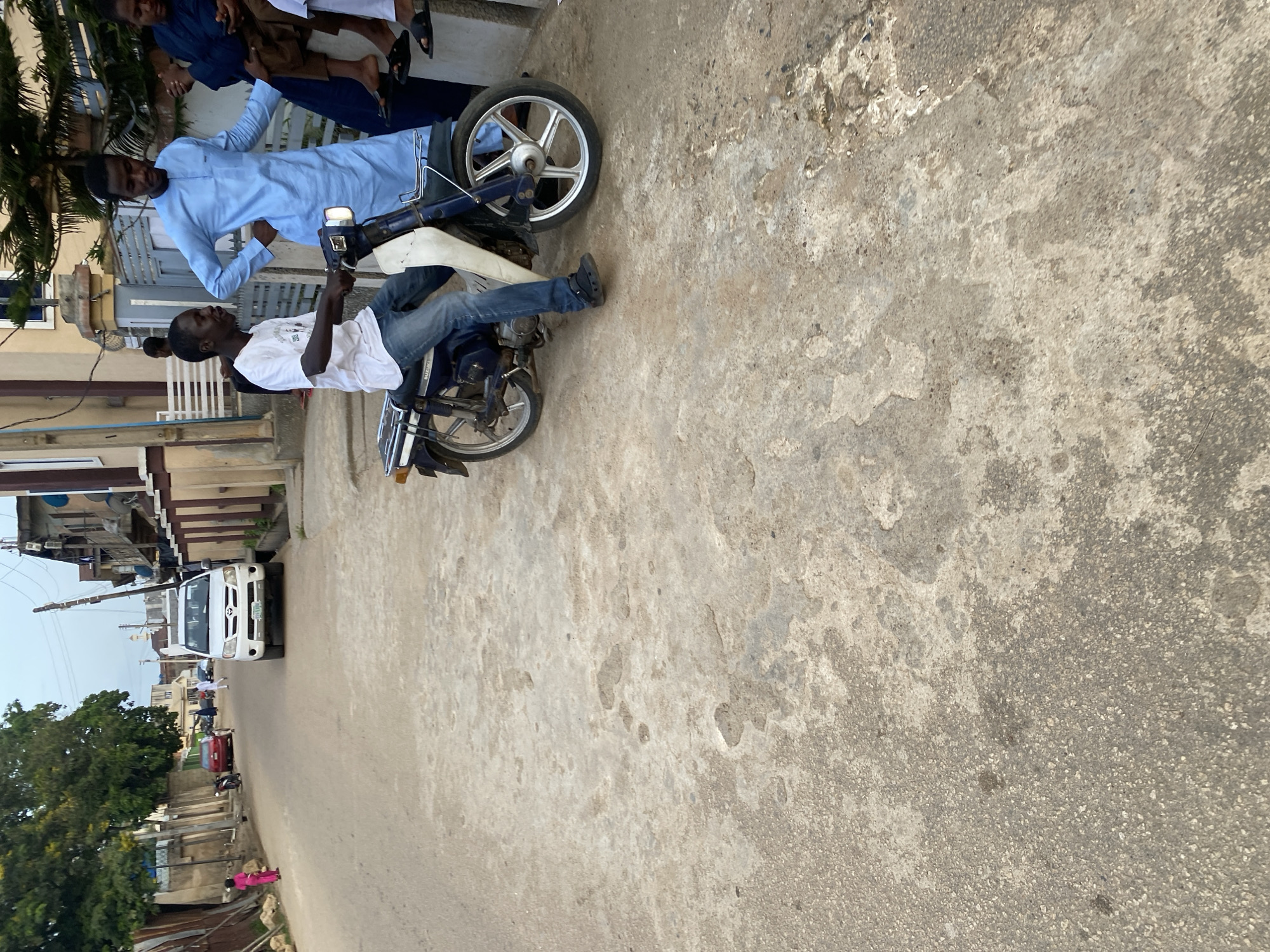 A man in a white t-shirt and blue jeans is seated on a blue motorcycle, holding the handlebars. Beside him, a man in a light blue robe stands, facing left. The scene takes place on a paved street in Jos, Nigeria, with buildings and trees lining the background. A white SUV is parked in the distance on the left side of the street, with a few other figures and a red car also visible. The ground is a rough, light-colored asphalt or concrete. The lighting suggests it is daytime, possibly late afternoon.