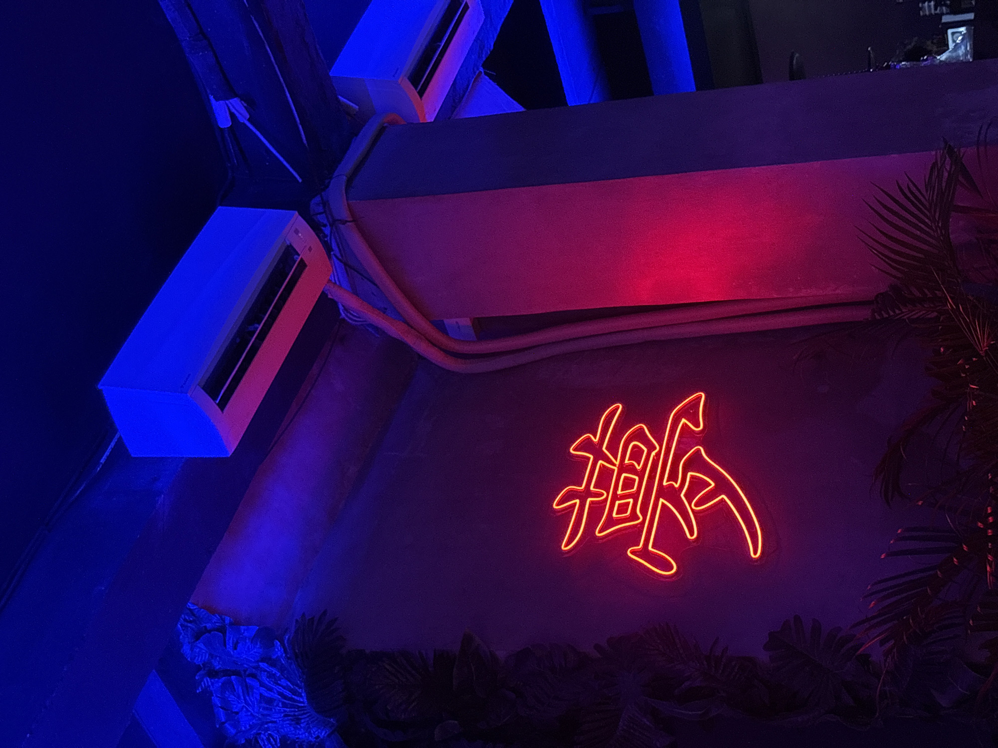 The image shows a dimly lit interior, likely a bar or restaurant, in Kuta Selatan, Indonesia. The walls are decorated with tropical foliage and a large, glowing neon sign in the shape of stylized Asian characters. Air conditioning units are visible mounted on the ceiling. The lighting is dominated by blue and red hues. There are no people visible.