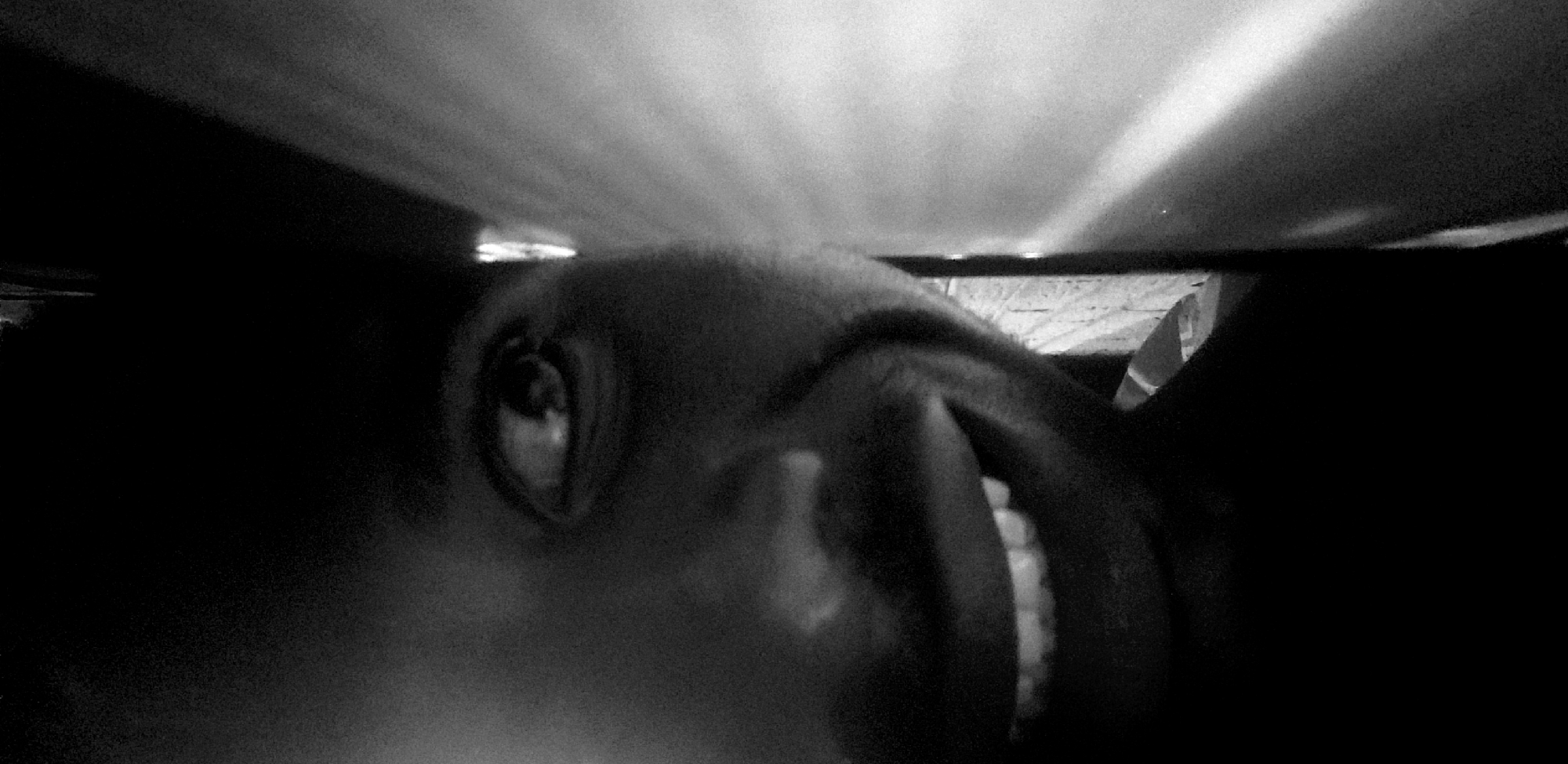 The image shows a close-up, low-angle view of a person's face in black and white. Only one eye, part of the nose, and a section of the mouth showing teeth are clearly visible. The person appears to be lying down, with the camera positioned very close to their face. The background is dark, with some areas that seem to be illuminated by a light source above. The location in the city of Narayi, Nigeria is not apparent from the image.