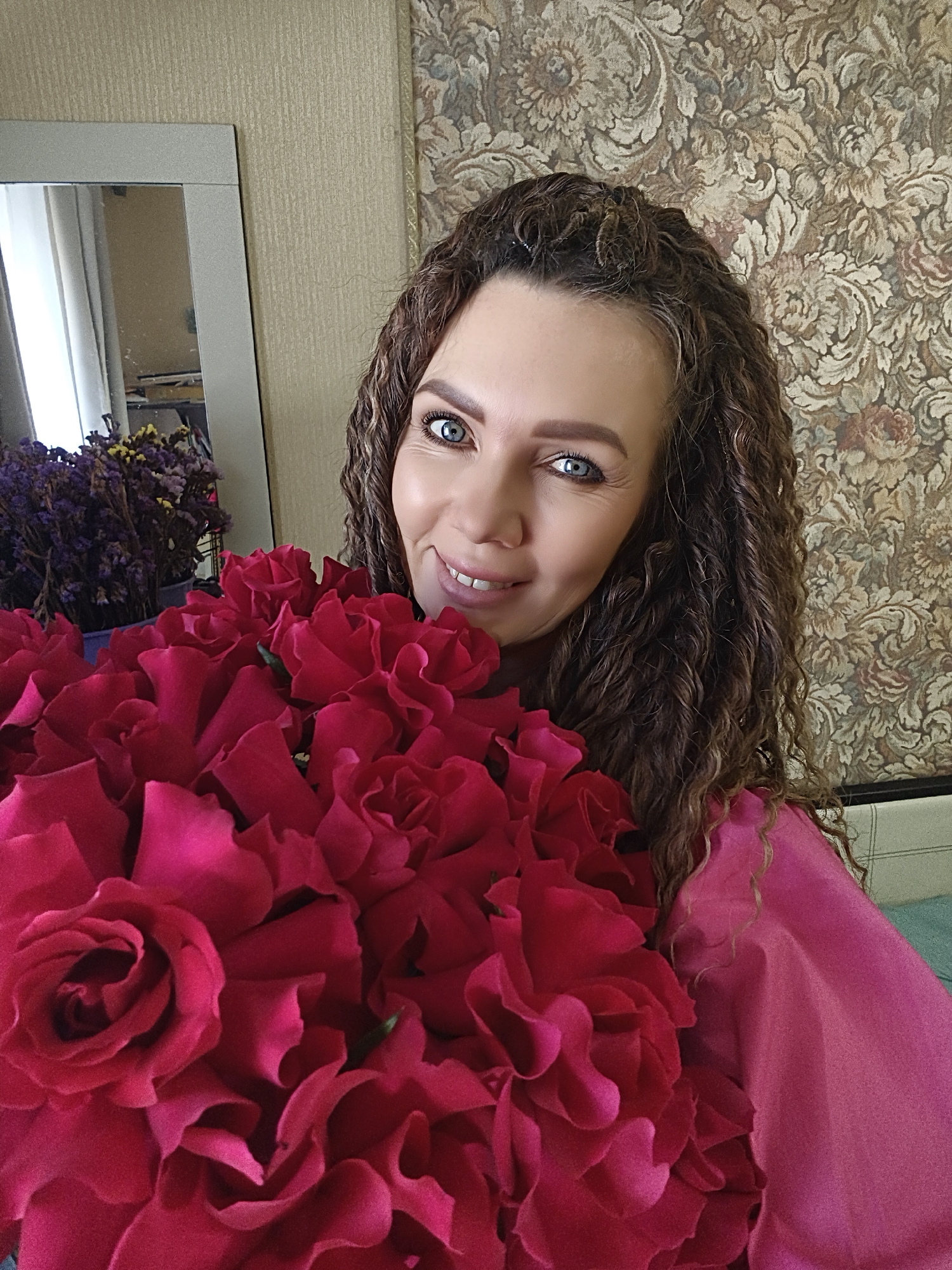 A woman with curly brown hair and blue eyes is holding a large bouquet of red roses. She is wearing a pink shirt. Behind her is a patterned wallpaper and a mirror reflecting another bouquet of flowers. The location is in the city of Unknown, Unknown.
